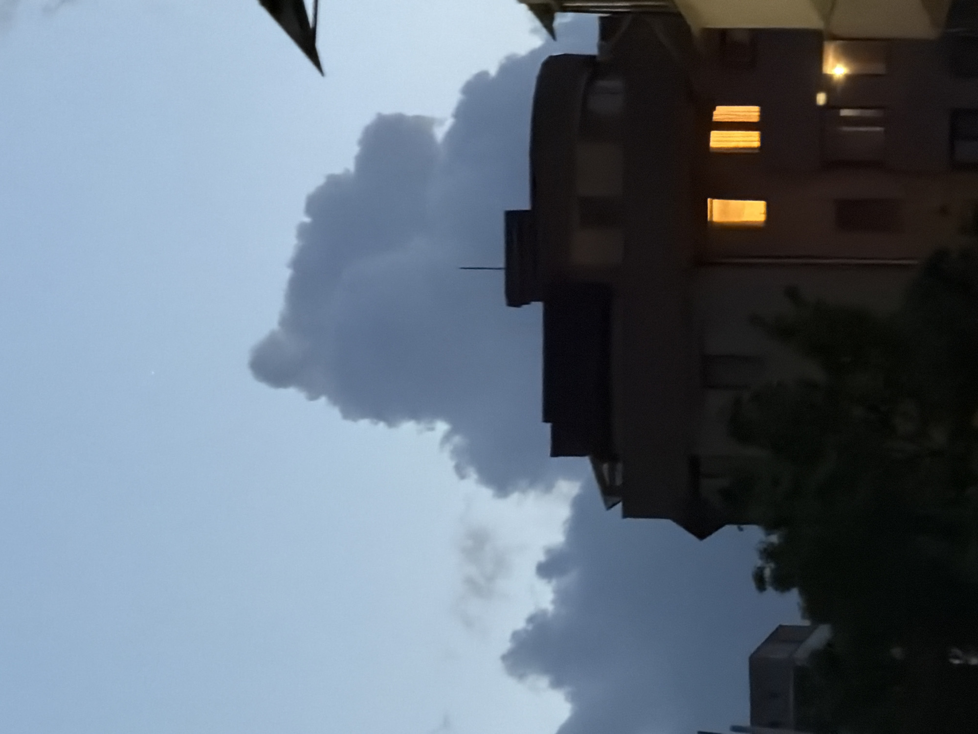 The image shows a view of the sky with dark clouds forming behind a building. There are a few visible lit windows in the building. The location is in the city of Sofia, Bulgaria, based on visual features and known context. A tree is partially visible in the foreground, and other buildings are visible in the background.