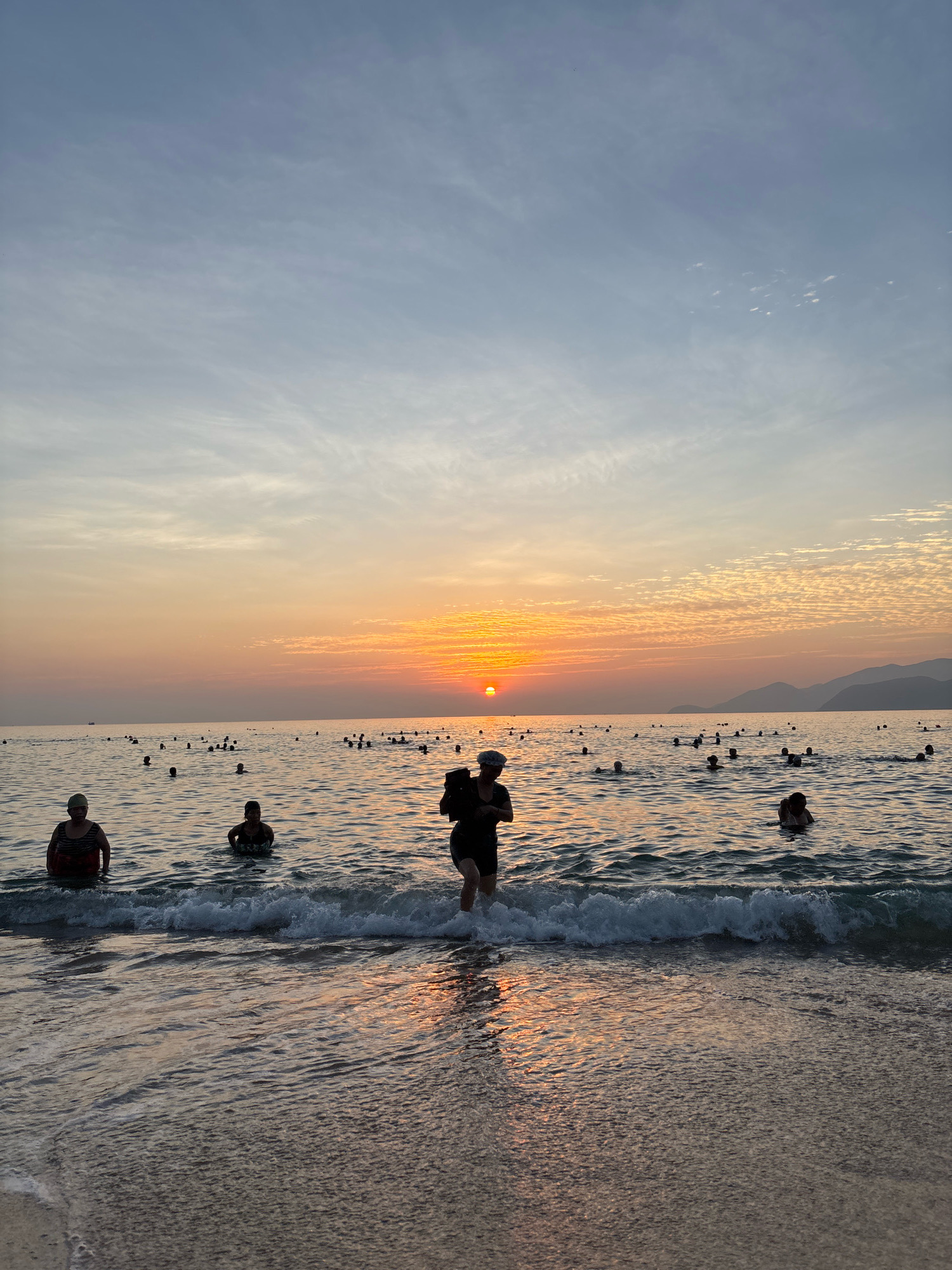 A wide shot captures a beach in Nha Trang, Vietnam, during a sunset. The sun, a bright orange orb, is low on the horizon, casting a warm glow across the sky and reflecting on the rippling water. The sky is a gradient of soft blues and oranges, with scattered wispy clouds. The sea is populated with numerous people wading and swimming. In the foreground, three individuals are visible closer to the shore. One person, wearing a dark swimsuit and a light-colored hat, stands in the surf. Another person in the middle ground is partially submerged, facing away from the camera. To the left, a person in a striped swimsuit and a cap is also in the water. The shoreline is a gentle slope of sand with small waves breaking. In the distance, a line of mountains is silhouetted against the twilight sky.
