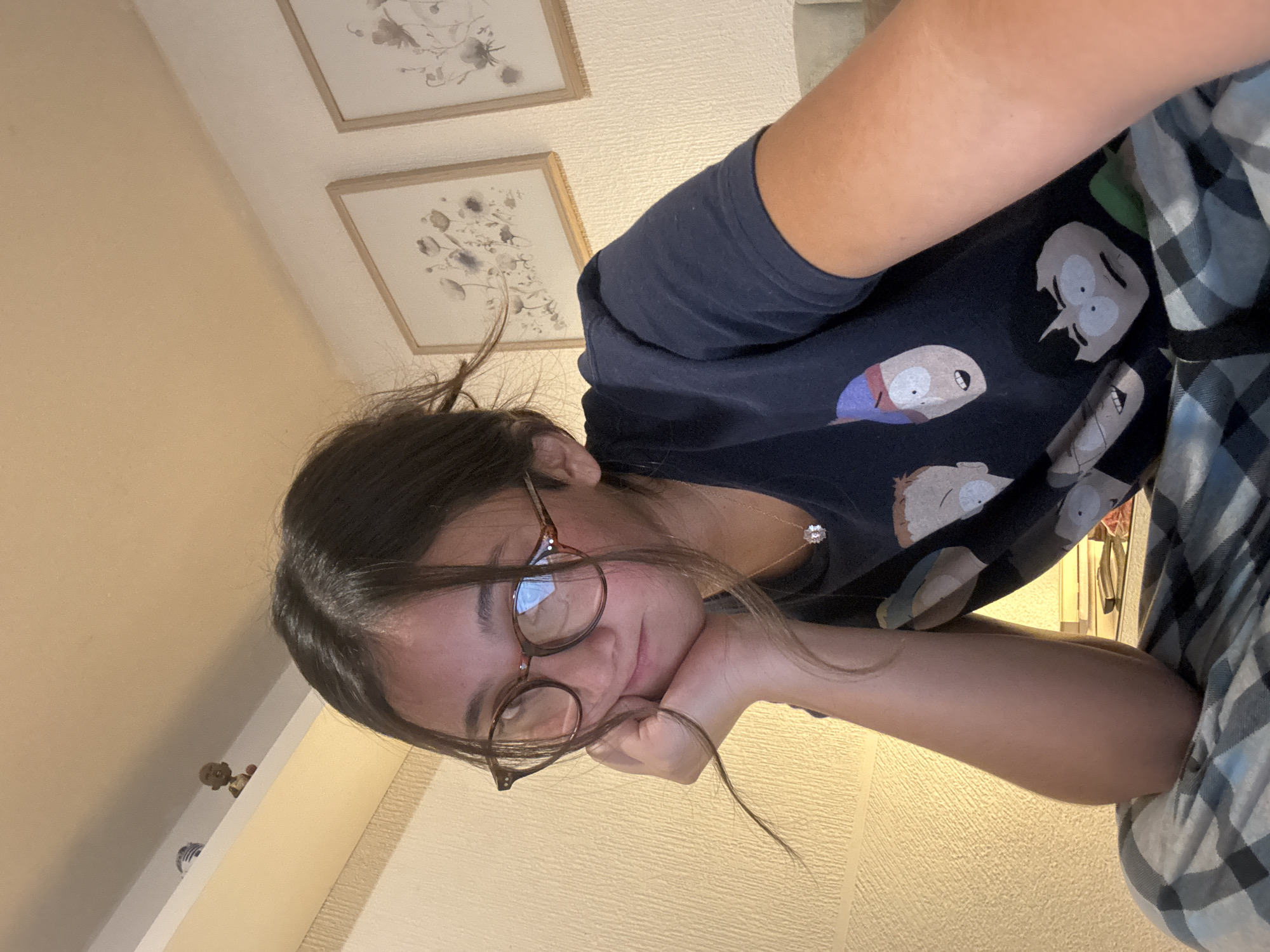 A young woman is lying down indoors, possibly on a bed, with her head propped on her hand. She is wearing glasses, a dark t-shirt with cartoon characters on it, and patterned pajama pants. Behind her are two framed pictures on the wall. A shelf with figurines is visible in the background. The location is not verifiable as San Bernardino Tlaxcalancingo, Mexico with only the given information.