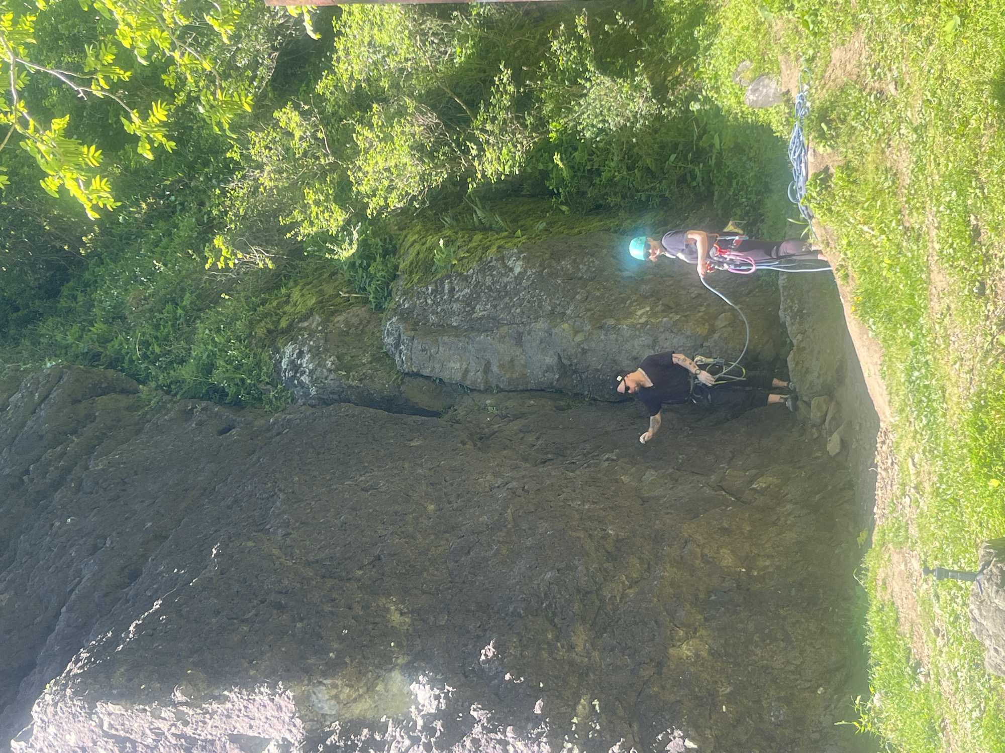 Two individuals are engaged in rock climbing at a location characterized by a tall, gray rock face overgrown with vegetation. A person wearing a black shirt, black pants, and a black hat is actively climbing the rock. Above them, a second person, wearing a blue helmet, a black shirt, and black pants, is situated on a ledge, presumably belaying. Ropes connect both individuals. The surrounding environment includes dense green vegetation and grassy areas. The location is consistent with a natural rock climbing area, potentially outside a city. The available image and context do not provide sufficient information to confirm that the location is in Batumi, Georgia.