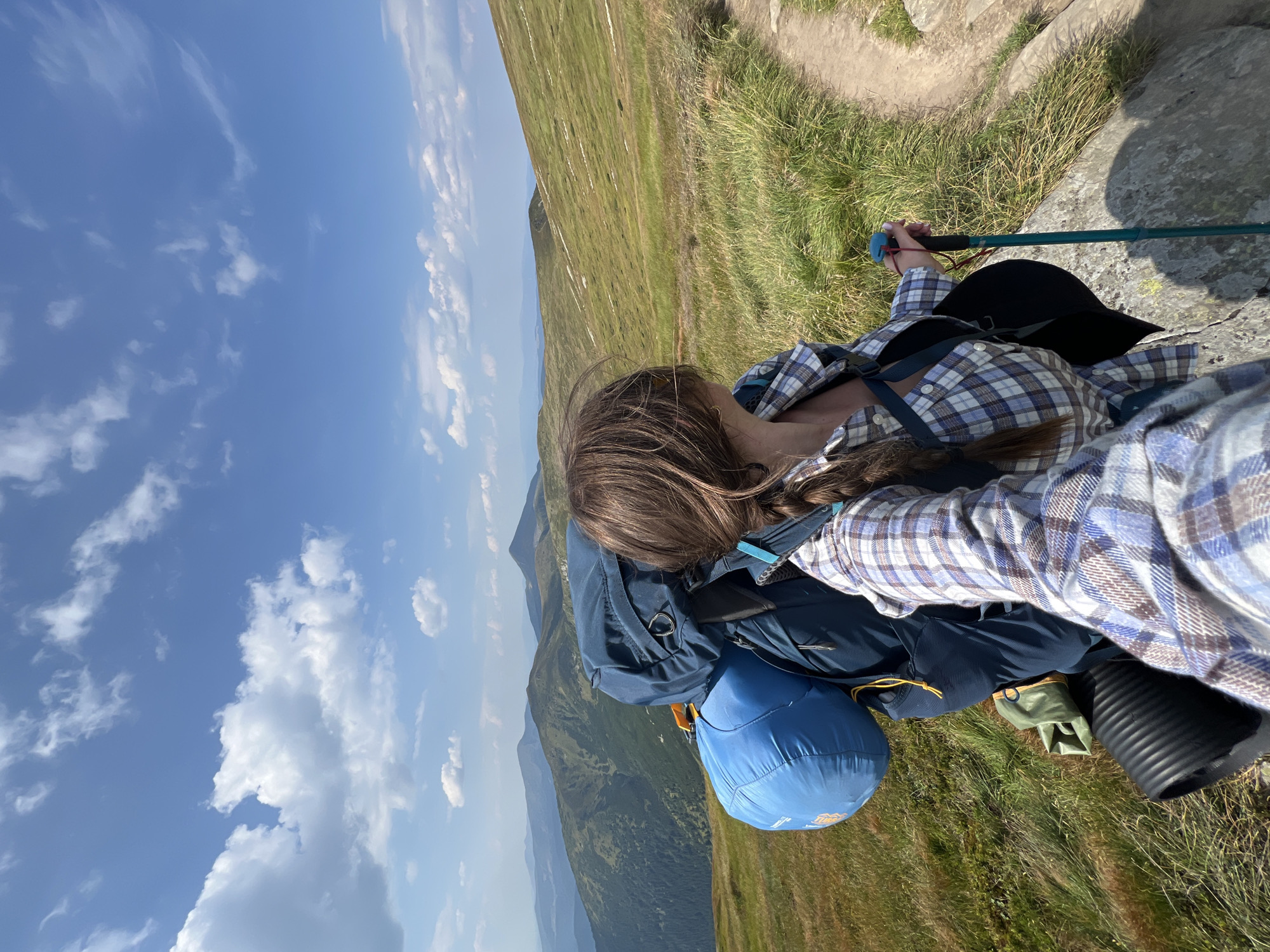 A woman is hiking in the Carpathian Mountains near Hoverla, Ukraine. She is wearing a plaid shirt and a backpack, and she is holding a hiking pole. The landscape is mountainous, with green hills and a blue sky with white clouds.