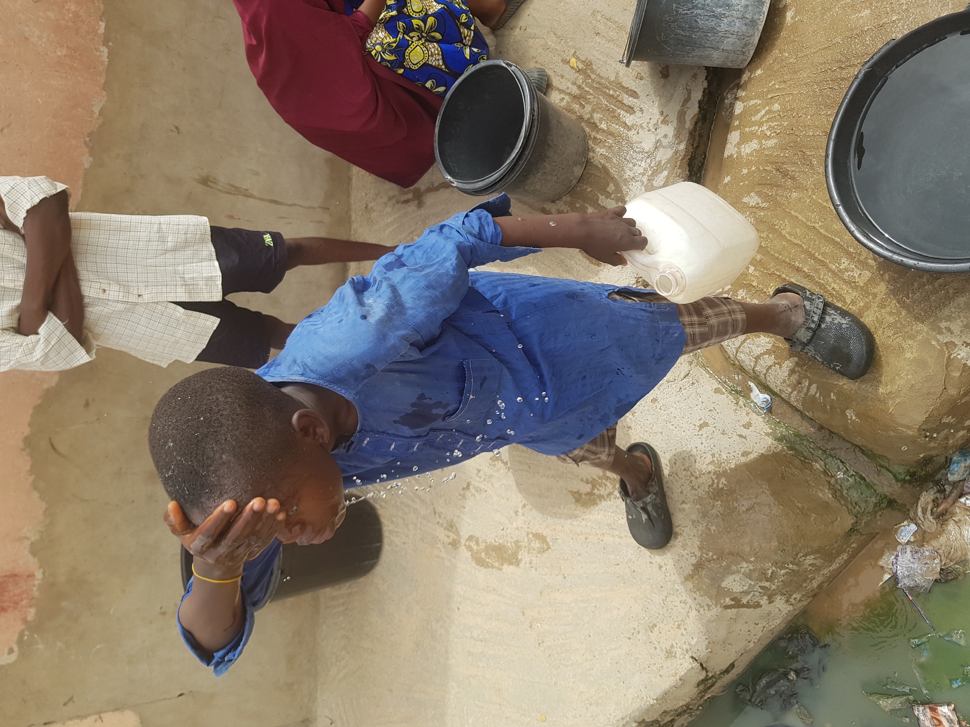 An overhead shot shows a dark-skinned person in a blue shirt and checkered shorts washing their face with water from a white plastic jug. They wear dark, slip-on shoes. Water pours from the jug over their face and onto the concrete ground. To the left, a person in a tan and white shirt with dark shorts stands nearby. Behind the person washing their face, a woman in a red dress with a blue and yellow floral pattern sits next to a gray bucket. Another gray bucket sits nearby, and a black basin filled with water is to the right. The scene is set on a concrete surface near a drain filled with greenish water and trash. This image was taken in Dutsinma B, Nigeria.