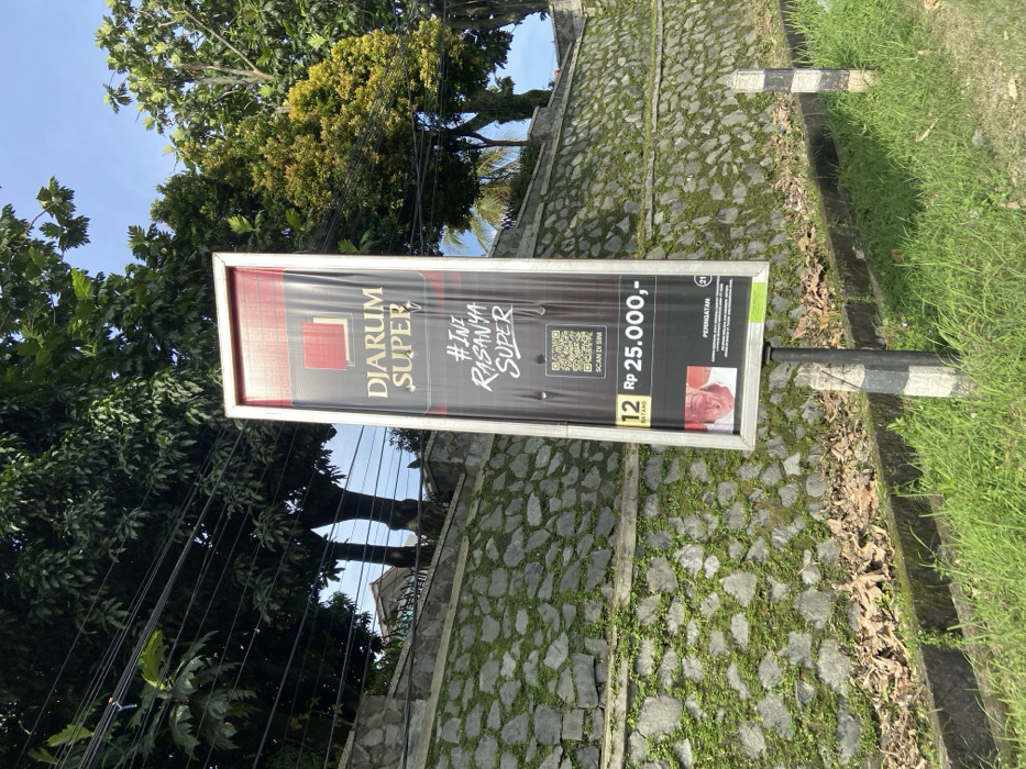 A large, rectangular, silver-framed advertisement sign is mounted on a black pole next to a stone wall covered in green moss and vegetation. The advertisement features a red and white logo, "DIARUM SUPER," and the tagline "#INI RASANYA SUPER," displayed on a white background. Below the logo is a QR code labeled "SCAN DI SINI," indicating that it is a scannable code. The sign also includes a price, "Rp 25.000,-," and a quantity, "12 BATANG." A small image of a man's face appears at the bottom left corner of the advertisement, with the word "PERINGATAN" below it. The sign is set against a backdrop of lush green trees, a clear blue sky, and a few power lines overhead. The scene is located in Jatiluhur, Indonesia, at the coordinates -6.57 latitude and 107.438 longitude.