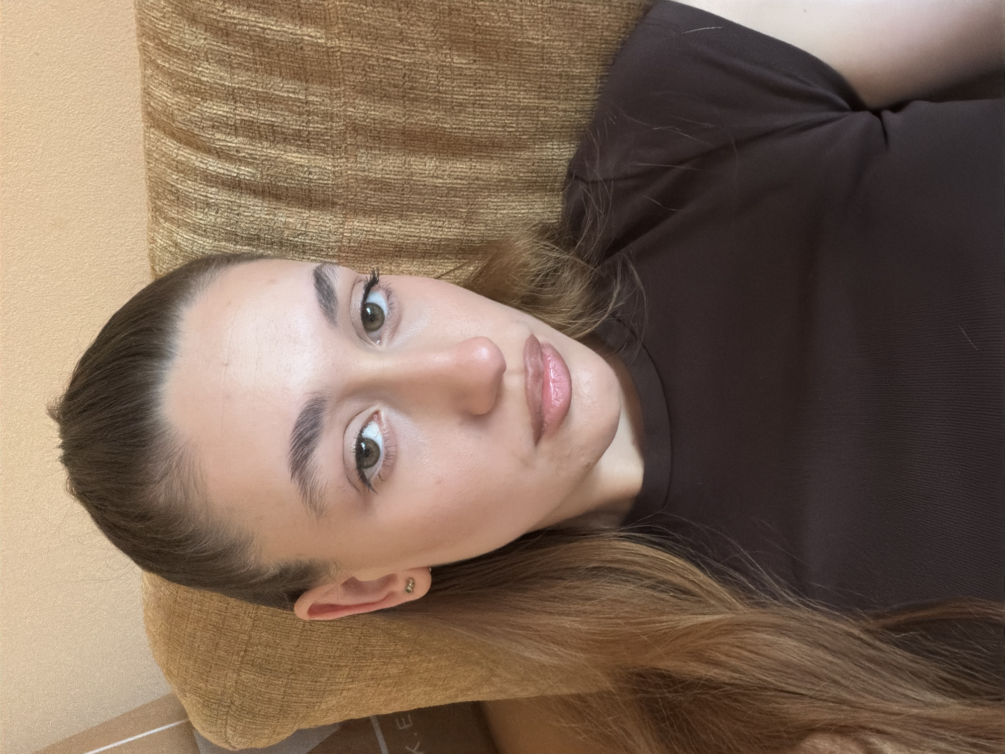 A woman lies on a tan couch with her head resting on a pillow. She has light skin, brown hair pulled back, and light eyes. She is wearing a dark brown, fitted shirt. The location in Plovdiv, Bulgaria cannot be determined from the image.