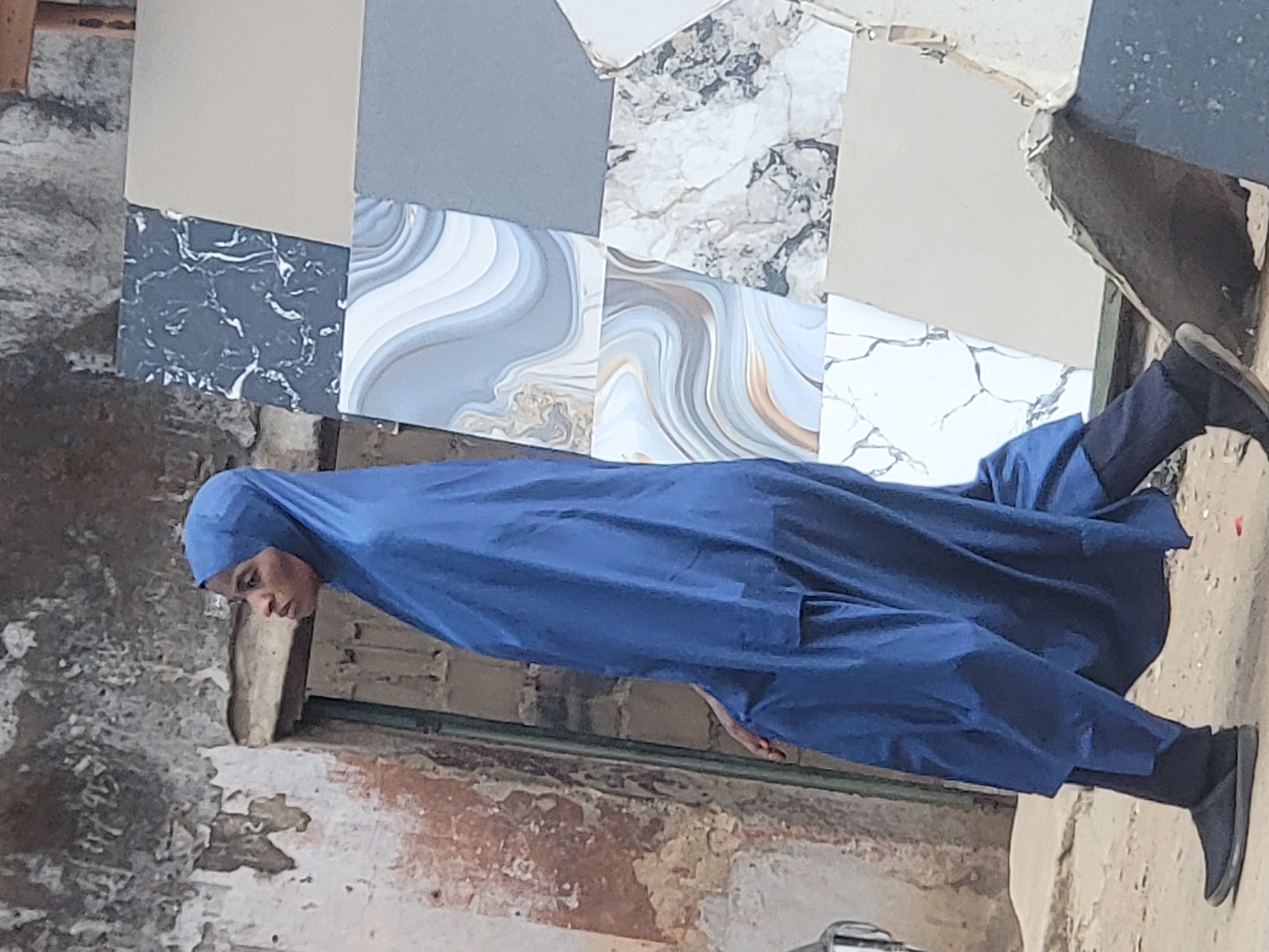 A woman wearing a blue burka stands in front of a wall with tile samples attached to it. The wall is cracked and shows signs of wear. The location is Maiburiji, Nigeria. The woman is facing towards the left of the frame. She is wearing black shoes and her head is covered.