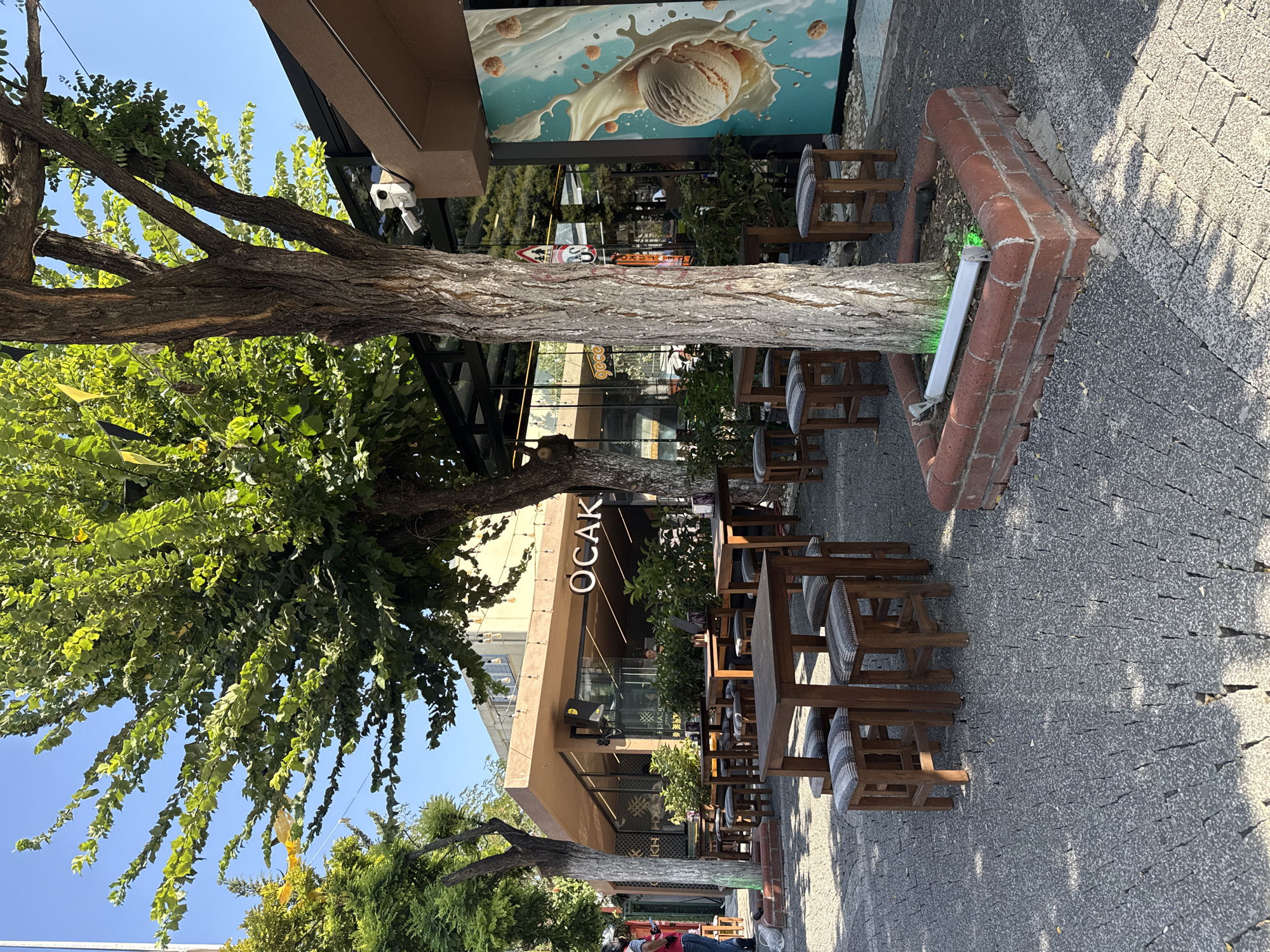 The image shows an outdoor seating area of a cafe named "OCAK" in an unknown city. Wooden tables and stools are arranged on a cobblestone pavement. Trees with green foliage provide shade. The cafe has large glass windows, and a mural of ice cream is visible on the side of the building. A security camera is mounted on the exterior wall. The scene appears to be captured during the daytime, with bright sunlight casting shadows. No people are actively engaged in any visible actions, though one person is partially visible on the far left of the image. The environment is clean and well-maintained.