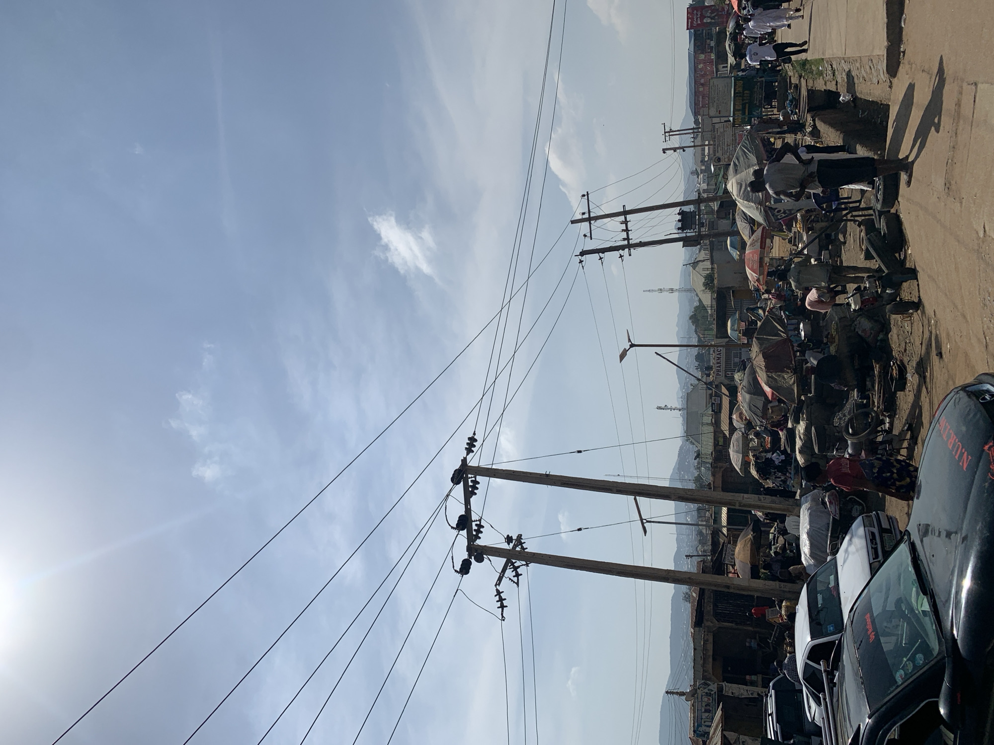The image depicts a street scene in Rafin Par, Nigeria. Several cars are parked on the right side of the frame, with a black car prominently positioned in the foreground. Along the road, a market area is visible, filled with people, goods, and makeshift stalls covered with umbrellas. Power lines and wooden utility poles run across the sky. In the background, there is a view of distant mountains partially obscured by haze.