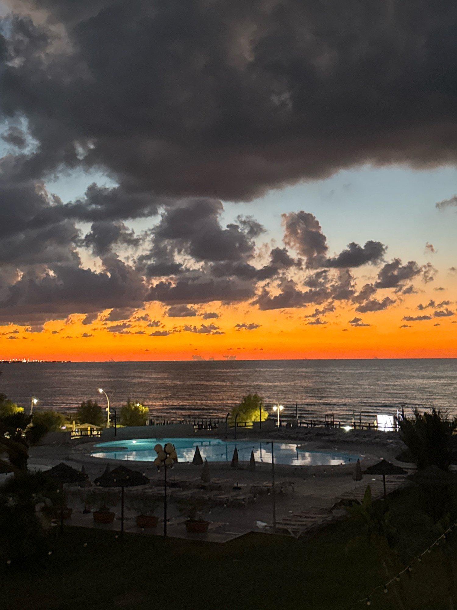 The image depicts a dusky scene at a seaside resort, likely in Isola delle Femmine, Italy, given the characteristic Mediterranean coastline. The sky is a dramatic spectacle of contrasts, with heavy, dark clouds dominating the upper portion, pierced by streaks of vibrant orange and yellow from the setting sun near the horizon. A vast expanse of the sea stretches across the middle ground, its surface reflecting the colors of the sky and appearing somewhat choppy.
In the foreground, a swimming pool area is visible. The pool itself is illuminated, casting a blueish glow onto its surface and the surrounding pavement. Several lounge chairs and umbrellas are scattered around the pool deck, suggesting a recreational area. The landscaping includes some potted plants and trees, as well as a grassy lawn in the very front. The presence of streetlights and possibly some distant city lights along the coastline indicate human habitation or development. No people are clearly discernible in the image, implying the resort is either closed for the evening or unoccupied at the moment of capture. The overall atmosphere is one of twilight, with natural light fading and artificial lights beginning to assert themselves.