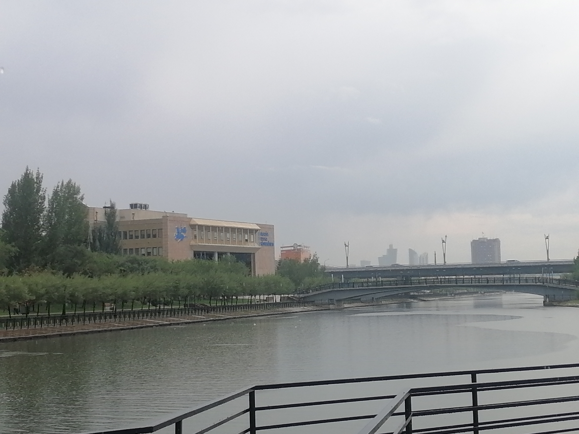 In the city of Unknown, Unknown, a wide river flows through a cityscape under a cloudy sky. On the left bank of the river, a modern building with a light-colored facade and blue lettering is visible, flanked by rows of green trees. Beyond the trees, other buildings and structures can be seen in the distance. A bridge with a pedestrian walkway and vehicular lanes crosses the river. The water is calm, reflecting the overcast sky. In the foreground, a dark metal railing of a walkway or balcony is visible.