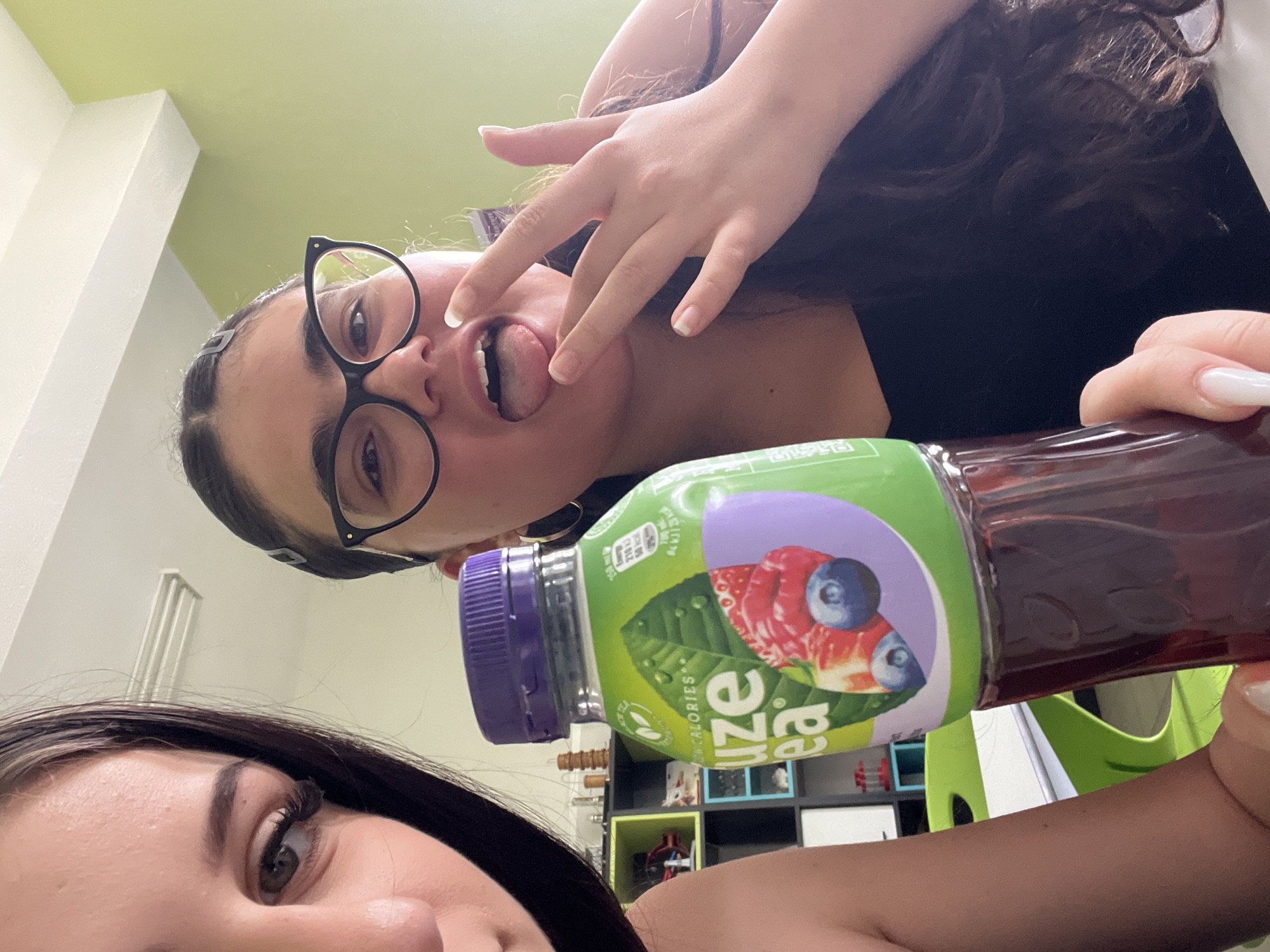 Two women pose in a selfie. One woman, wearing black-rimmed glasses and dark hair, sticks her tongue out, touching it with her fingers. The other woman's face is partially visible in the lower portion of the frame. A bottle of Lipton Ice Tea is being held in front of them. The location is indiscernible; there is no evidence to place this image in Plovdiv, Bulgaria.