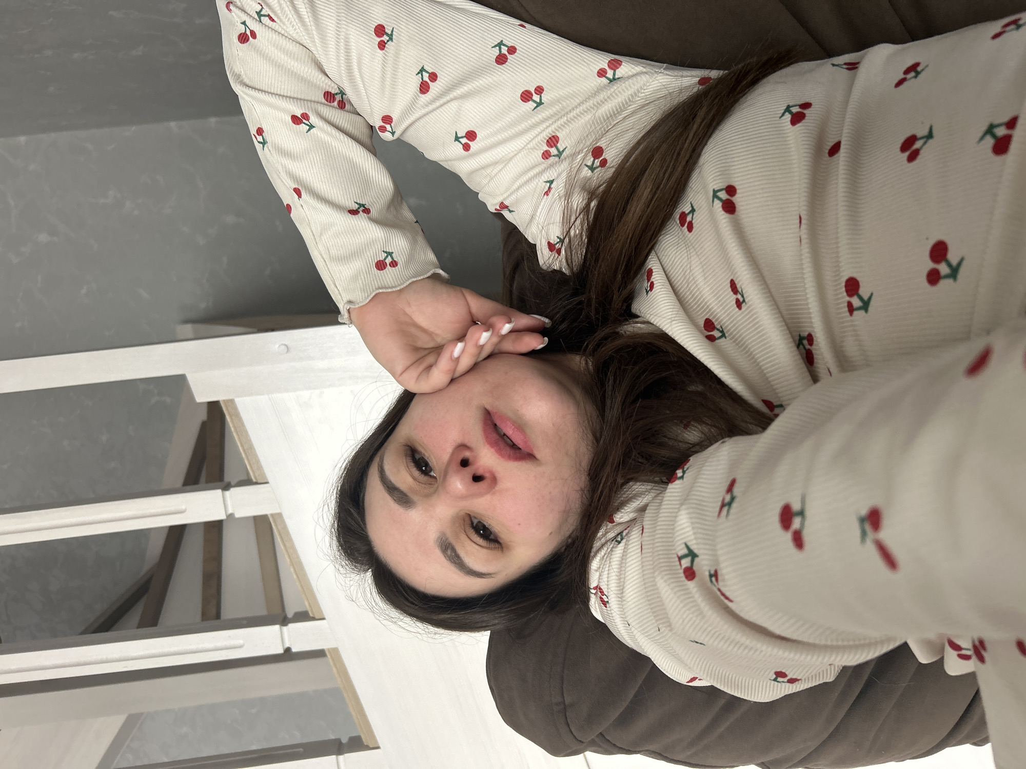 The image shows a woman lying on a brown upholstered surface, possibly a couch or bed. She is wearing a white, long-sleeved shirt with a cherry pattern. Her right hand is near her face. Her hair is dark brown. The background includes a gray and white wall and a white shelving unit. The location is Chaikovsky, Russia.