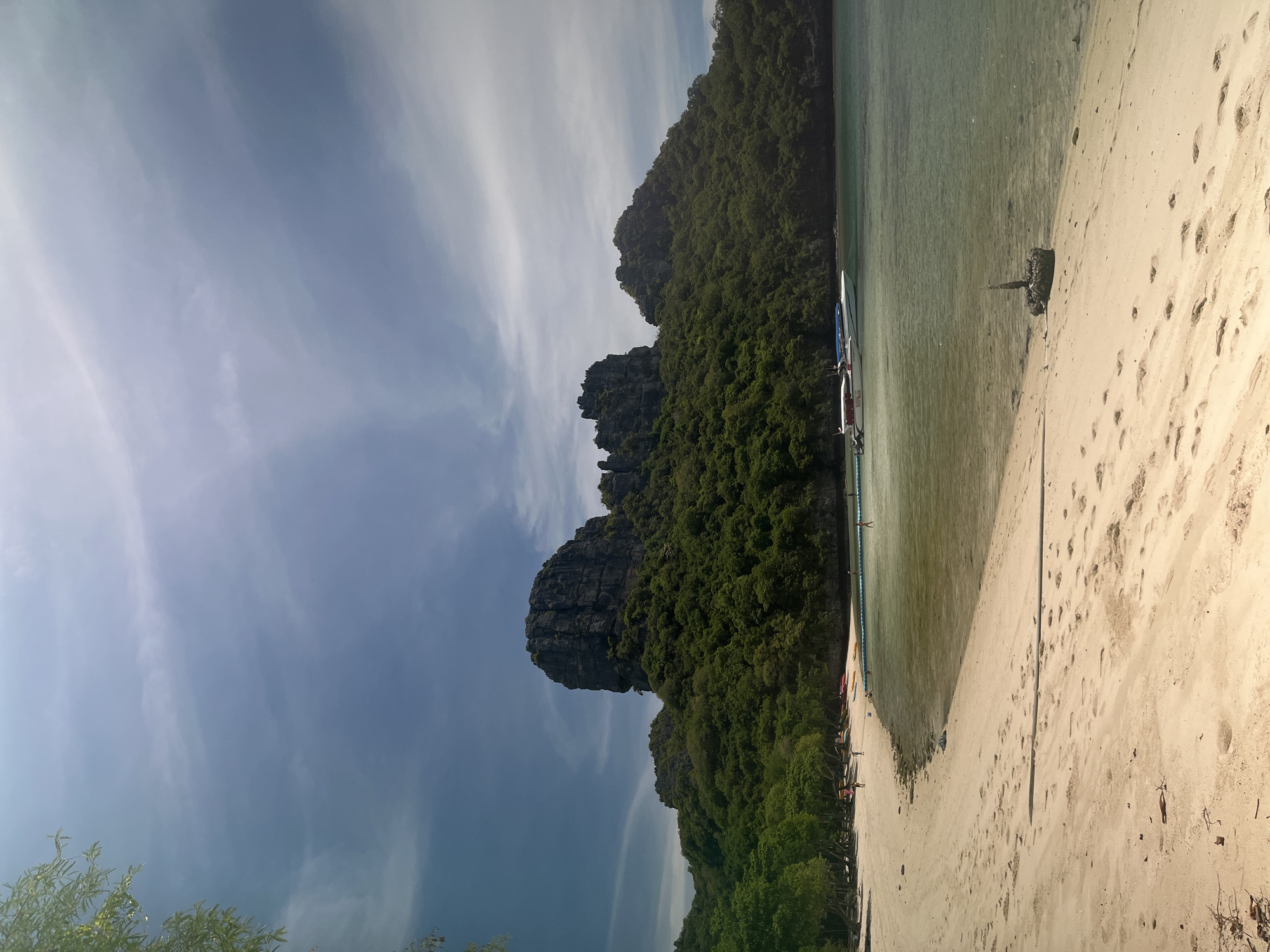 This image captures a serene tropical beach scene in Thailand. The foreground is dominated by a wide expanse of light-colored sand, marked with numerous footprints and a few scattered leaves and twigs. The sand slopes gently down to the clear, shallow water, which has a greenish hue. A dark, barnacle-encrusted object resembling a mooring or anchor is partially submerged at the water's edge on the right side of the frame.
In the middle ground, the water is calm and transparent, revealing the sandy seabed beneath. A long, blue floating barrier marks a boundary in the water. Two speedboats are moored along this barrier; one is white with red accents and a blue canopy, while the other is mostly obscured by the first, appearing to have a blue canopy. Several small, colorful kayaks are pulled up on the sand to the left of the boats.
A narrow strip of beach lines the edge of the water, populated by a few distant figures, some of whom appear to be walking along the shore or wading in the shallows. Behind the beach, lush green trees and vegetation form a dense backdrop.
The background is dominated by a large, imposing rocky hill covered in thick green foliage. The rock formations are jagged and steep, rising dramatically from the treeline. The sky above is a pale blue, with wispy white clouds stretched across it, suggesting a partly cloudy day. The overall impression is one of a beautiful and tranquil coastal landscape in a tropical setting.