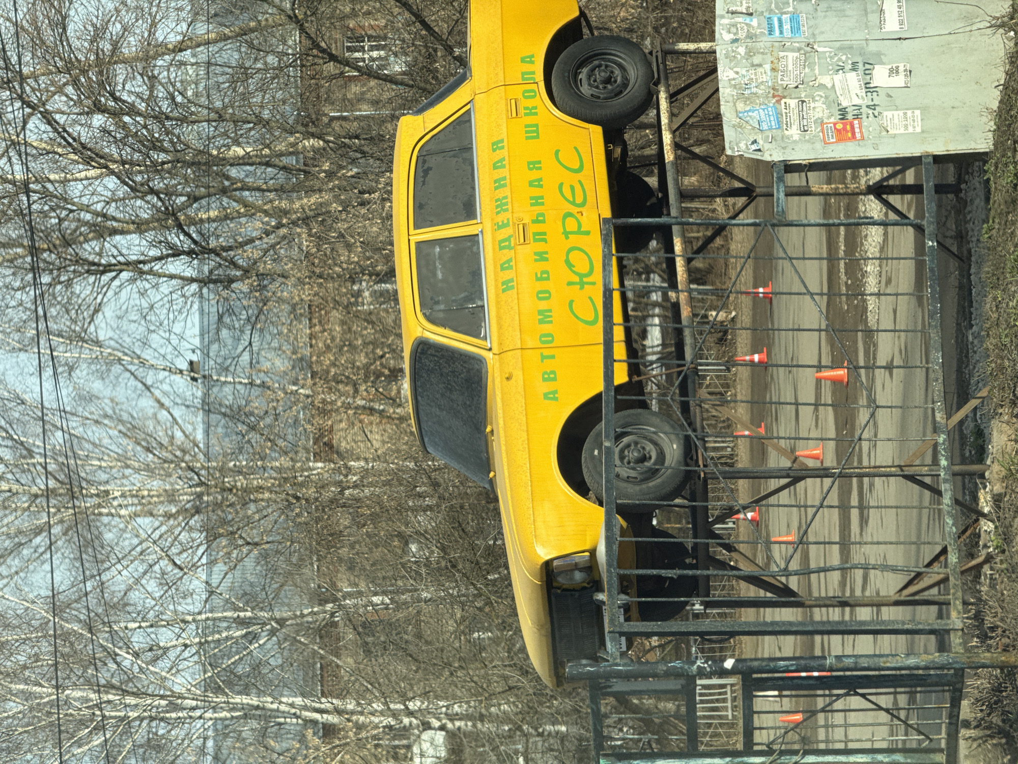 The image shows a yellow car mounted on a metal structure with the words "НАДЁЖНАЯ АВТОМОБИЛЬНАЯ ШКОЛА СЮРЕС" in green lettering, which translates to "Reliable Driving School Syures" in Russian. The background includes bare trees, a building with a window, and a metal fence with several orange cones placed along the roadway. There is a metal board with various posted flyers and job advertisements visible on the right side of the frame. According to OCR, the image includes information about work (РAБOTA) including contact numbers. Based on the Cyrillic text and other details, the location is likely Izhevsk, Russia.