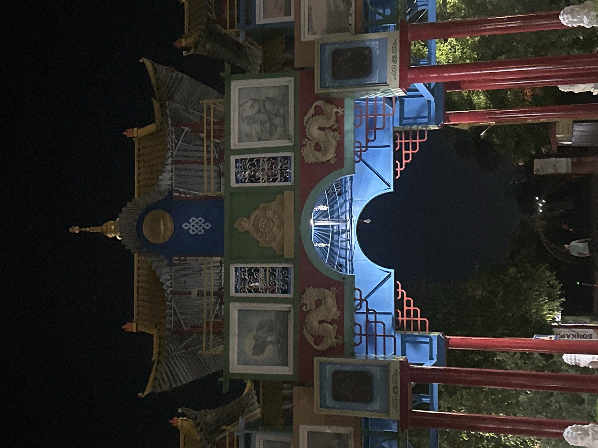 The image captures a night view of the Golden Gate (Zolotye Vorota), a prominent landmark in Elista, Russia, which is the capital of the Republic of Kalmykia, known for its Buddhist heritage. The gate is elaborately decorated with traditional Buddhist motifs and architectural elements. Red columns support a blue and gold structure adorned with paintings, sculptures, and intricate carvings. Golden dragons are depicted on the facade. The sky is dark, indicating nighttime, and the gate is illuminated, highlighting its vibrant colors and details. In the background, trees are visible, and a few people can be seen in the distance, walking through the archway. The overall impression is one of cultural and architectural significance, illuminated against the night sky.
