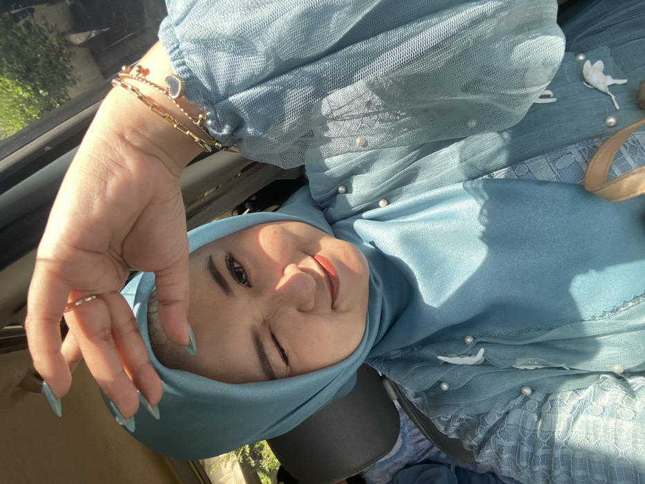 A female subject is photographed in a vehicle in Kebonagung, Indonesia, during daylight hours. She is depicted from the upper chest, wearing a light blue hijab and a matching layered garment featuring an inner textured fabric and an outer sheer tulle layer adorned with white pearls and white floral appliques. Her left eye is winking, and she displays a closed-mouth smile. Her lips are colored with a reddish-brown lipstick. Her right hand is raised, with light blue manicured fingernails, positioned above her head, potentially shielding from bright sunlight that illuminates her face from the upper right. Multiple gold-colored bracelets, including one with a dark heart-shaped charm, are present on her right wrist. A dark gray headrest is visible behind her left shoulder, and blurred outdoor greenery and a light-colored structure are visible through the vehicle window in the background.