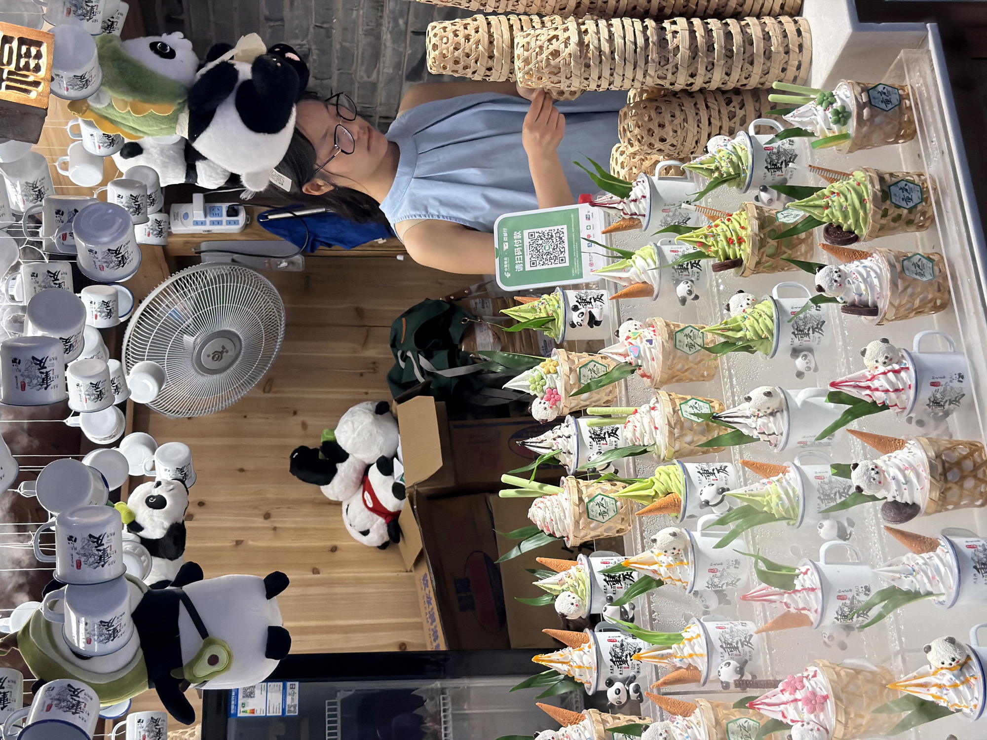 In Yuzhong District, China, a young woman with glasses, wearing a light blue sleeveless pleated top, stands behind a display of elaborately decorated soft-serve ice creams. The ice creams are presented in small mugs and woven baskets, topped with panda figures, colorful cones, and green leafy garnishes. Behind her, various panda plush toys are displayed, some hanging and some stacked. White mugs with black and red Chinese characters are suspended from the ceiling. A white oscillating fan is mounted on the wooden wall. To the right, stacks of woven baskets are visible. A QR code for mobile payment is displayed on a green sign in front of the woman. The overall scene suggests a food stall or shop specializing in panda-themed desserts.