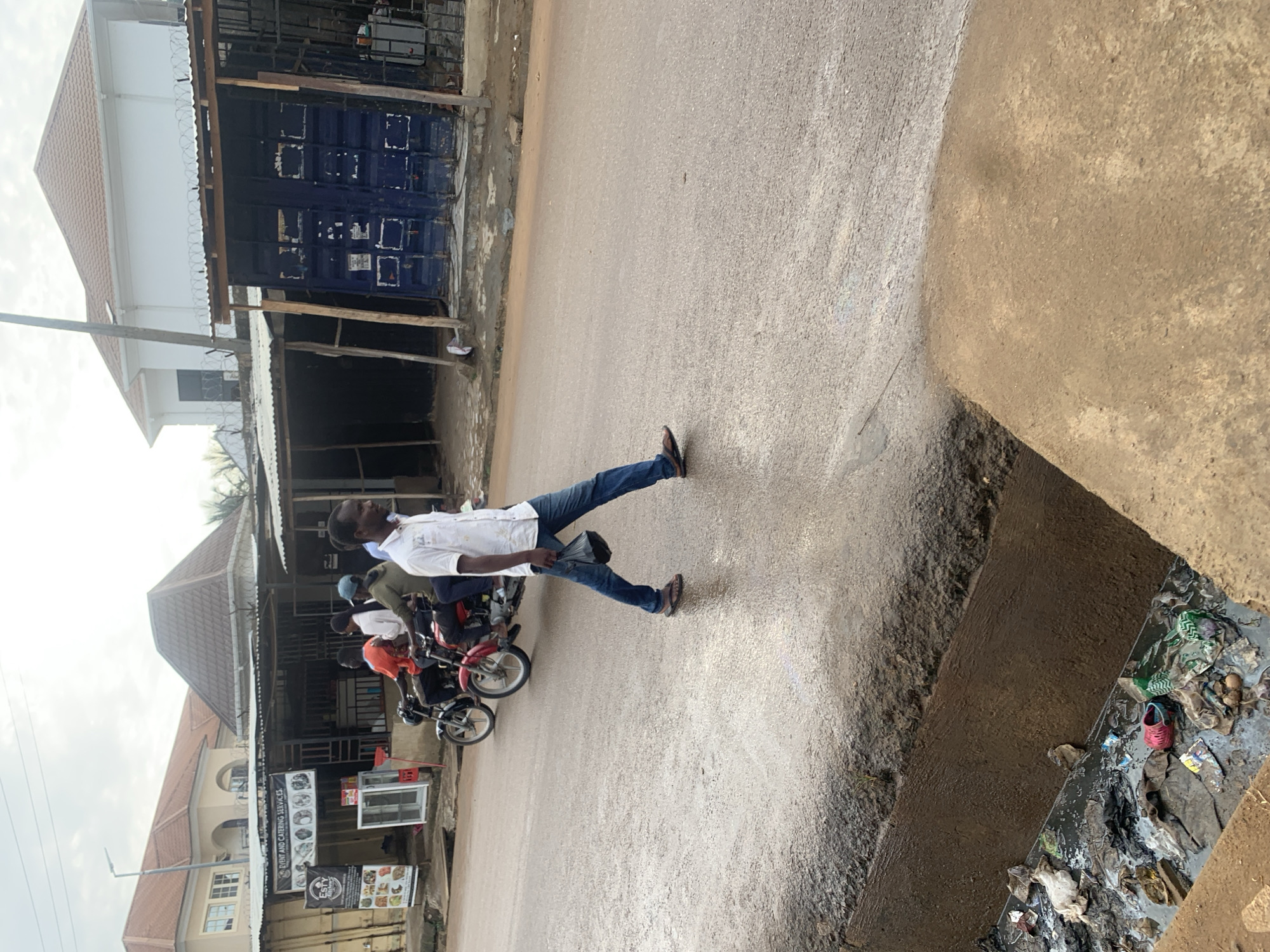 A man in a white short-sleeved shirt and blue jeans is walking across a wet asphalt road in Abuja, Nigeria. He is carrying a black plastic bag in his right hand and wearing brown flip-flops. In the background, two motorcycles are driving on the road. The first motorcycle, red, has two riders; the second motorcycle, black, has three riders. Behind the motorcycles, there are several shops and buildings. On the left, a building with a red-tiled roof and white facade is visible. In front of the shops, there are corrugated iron roofs. The road is lined with a concrete curb and a drainage ditch filled with trash. The sky is overcast.