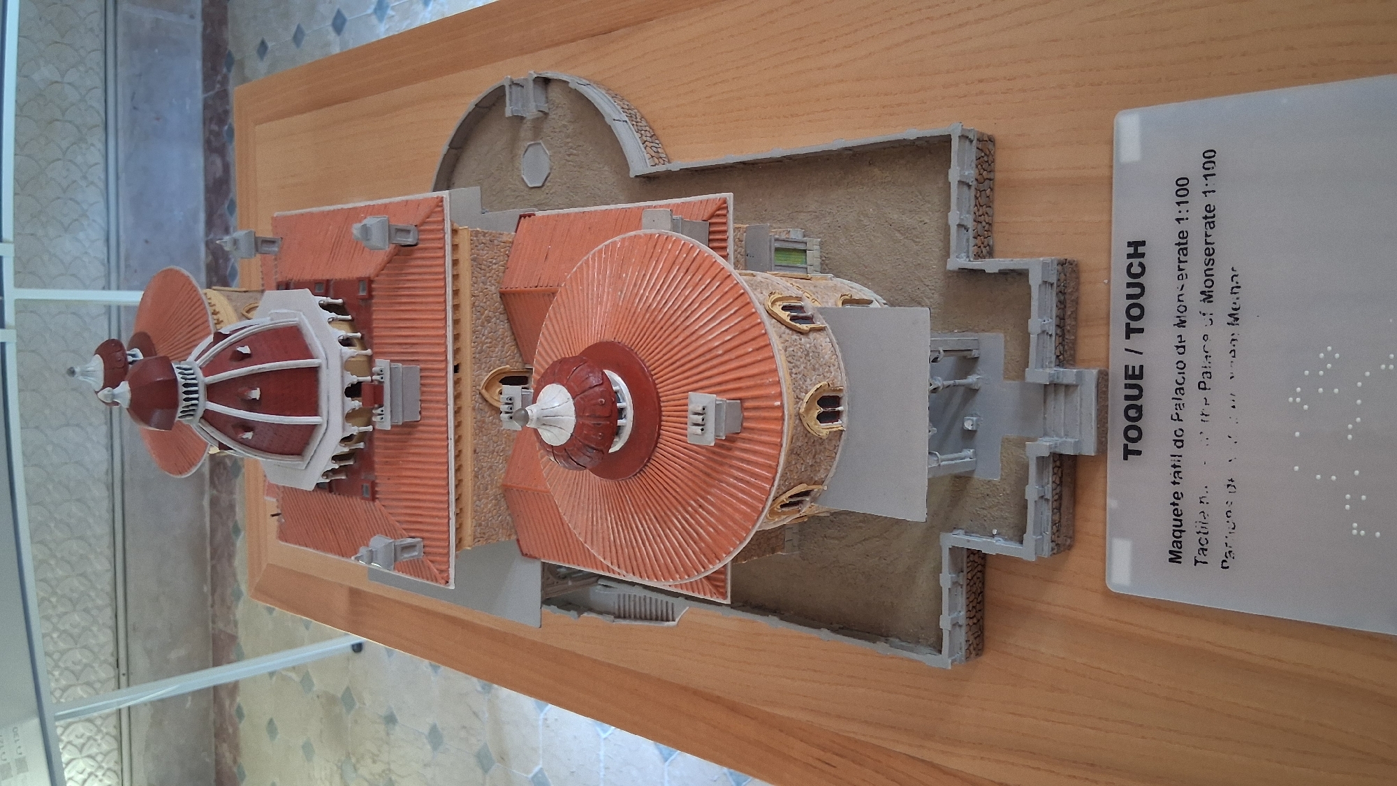 This is a top-down, slightly angled medium shot of a detailed architectural model of Montserrat Palace, located in Sintra, Portugal. The model, presented on a wooden table, features two main circular structures with conical, red-tiled roofs and prominent domes. The larger, rear structure has a white and red dome with architectural detailing, topped by a smaller finial. The front, smaller structure also has a red roof and a white and red dome with a finial. The model depicts the exterior of the palace, including stone walls, arched windows, a portico with columns, and staircases. The surrounding area of the model is made to resemble sand with miniature stone walls and pathways. In the foreground, a sign with text in Portuguese and English, along with Braille, provides information about the tactile model of Montserrat Palace at a scale of 1:100. The environment appears to be indoors, possibly a museum or exhibition space, with a patterned floor visible in the background and a white stand supporting a display above. There are no people visible in the image.