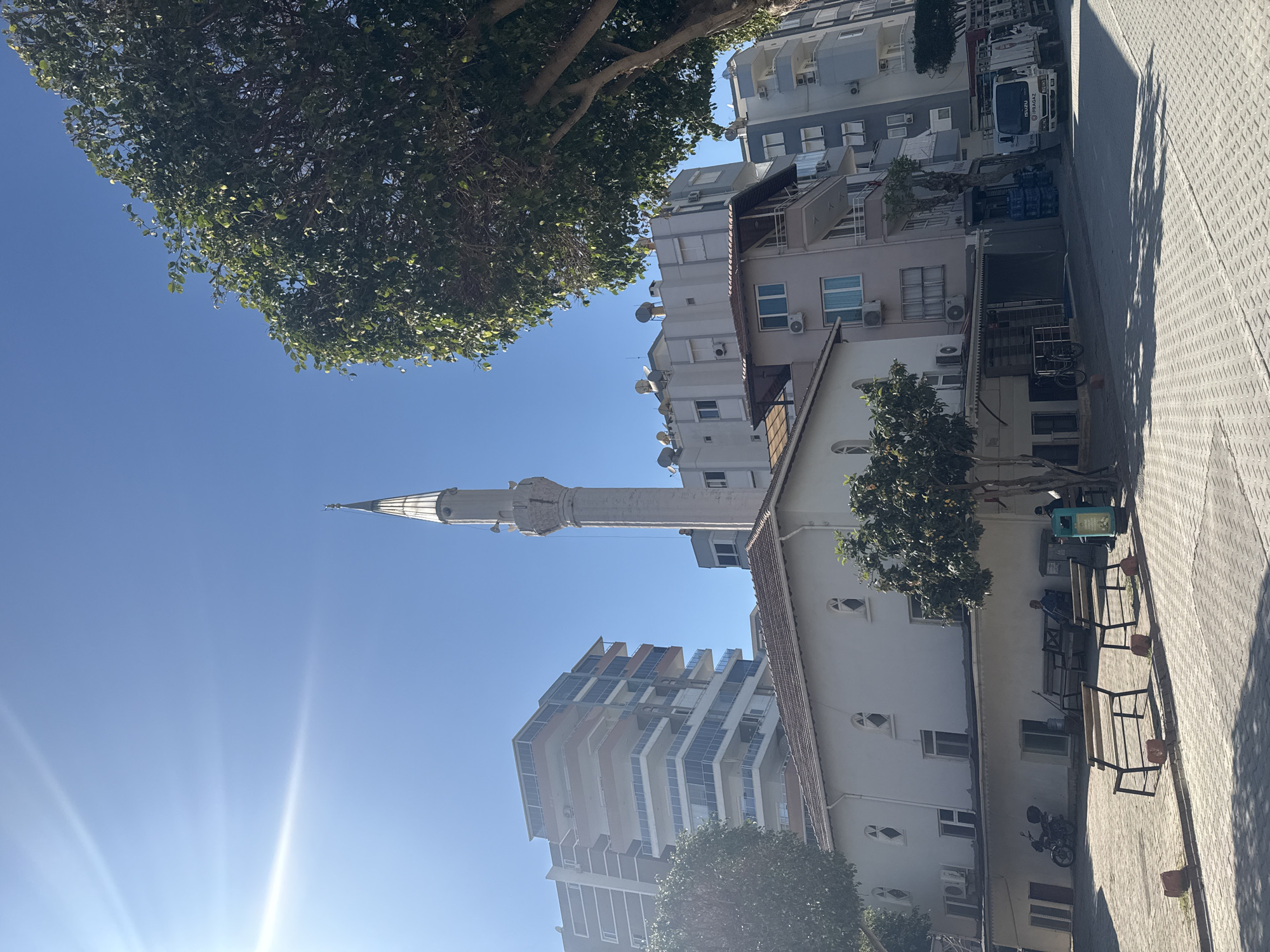 The image, taken in portrait orientation, captures a sunlit urban street scene dominated by a prominent minaret under a clear blue sky. A tall, slender white minaret with a conical roof and a small balcony near its top stands centrally, its pale structure contrasting with the deep blue.
To the left of the minaret, a modern multi-story building features numerous balconies and air conditioning units, its light-colored facade reflecting the sunlight. To the right, a more traditional building with a reddish-brown tiled roof and distinctive arched windows is partially visible, nestled amongst green trees. Another large tree with dense foliage frames the upper left corner of the scene.
On the right side of the image, a patterned sidewalk runs parallel to the buildings, showing long, sharp shadows cast by the intense sunlight, indicating a bright day. A white delivery truck with the text "PRAGAZ" and "19 KGZ" visible on its side is parked on the street. Nearby, street furniture including benches and a green public waste bin, as well as a parked motorcycle, are visible. The sun's rays are clearly emanating from the lower-left portion of the sky. The combination of the minaret and the visible text on the truck suggests a location in Turkey or a similar region.