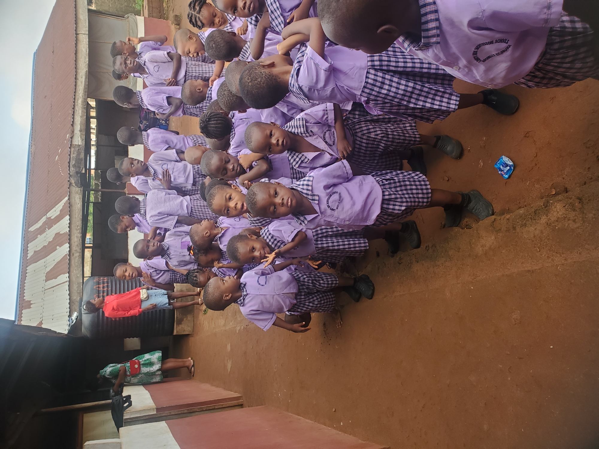 A group of children in purple school uniforms are assembled outdoors on a dirt ground, appearing to be at Liberty kid's Montessori School, Ukwu-Abwa, Nigeria. They stand in multiple rows with some facing forward and looking at the camera. The children all seem to wear black shoes. There are two adults visible near a building; one woman is wearing a green patterned dress and the other a red top and denim shorts. There is a water tank, and corrugated metal sheeting forming a roof. A crumpled blue wrapper lies on the ground amidst the children.