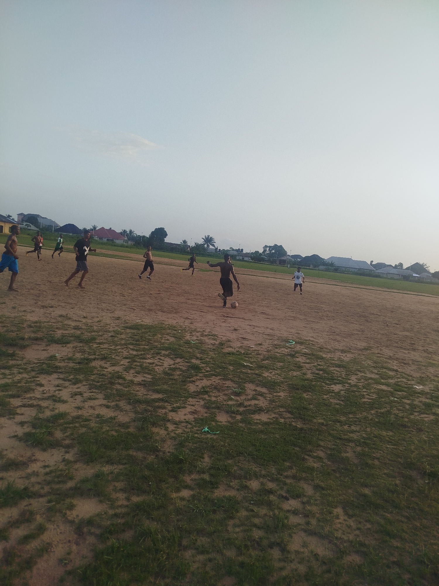 The image depicts a soccer game in progress on a dirt field in Ikot Mbonde, Nigeria. There are approximately 10 people visible, all male, actively involved in the game; some are running, and one is kicking a soccer ball. The environment is an open field with scattered grass patches, likely a local recreational area. In the background, there are houses and trees, indicating the edge of a residential area. The sky is overcast with hints of daylight.