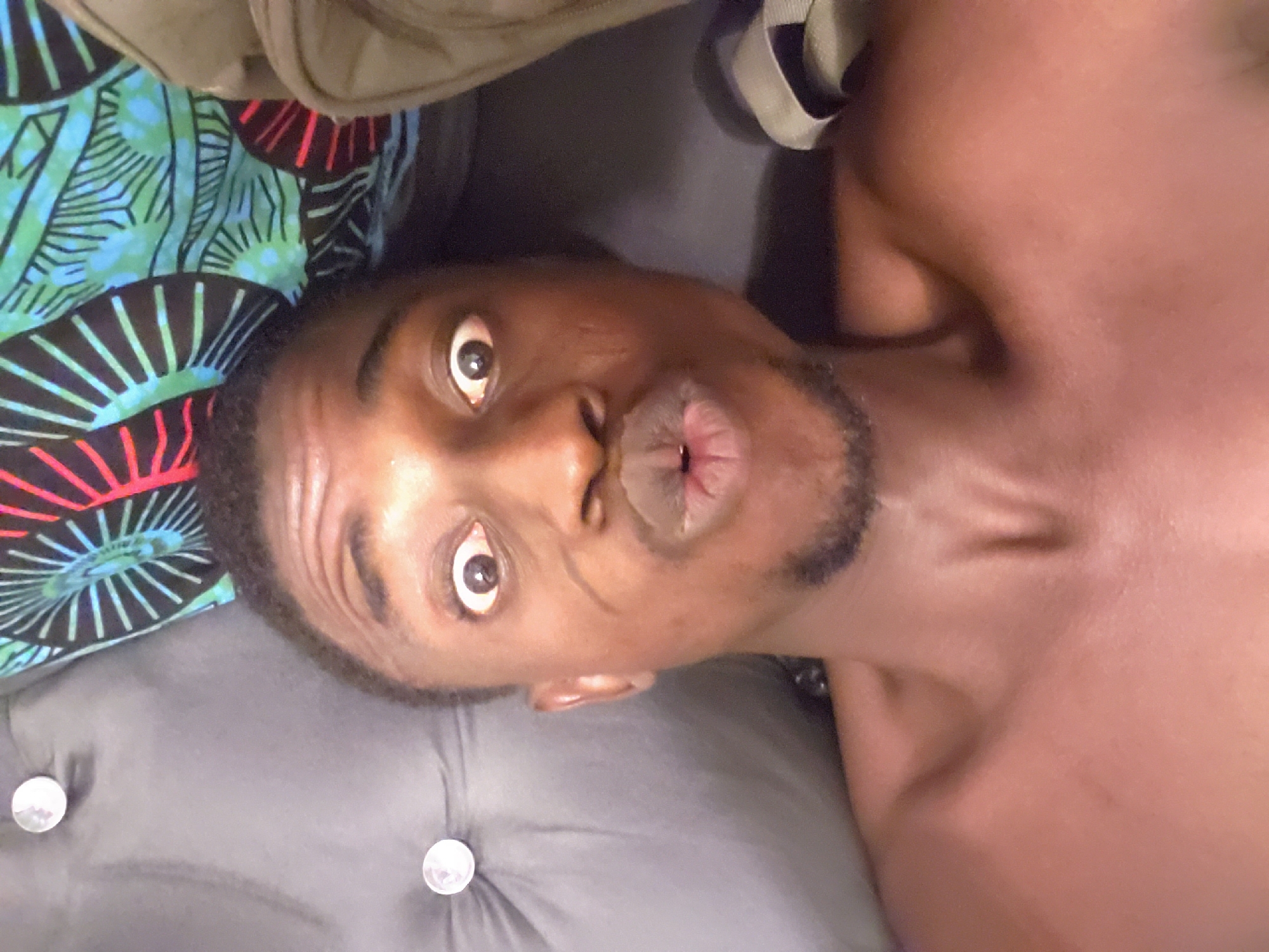 A close-up, upside-down selfie of a dark-skinned man with wide eyes and pursed lips. He has a short beard. Part of a patterned fabric and a gray, buttoned cushion are visible near his head. The location is unknown and cannot be verified as Adonkolo, Nigeria, based on the image alone.