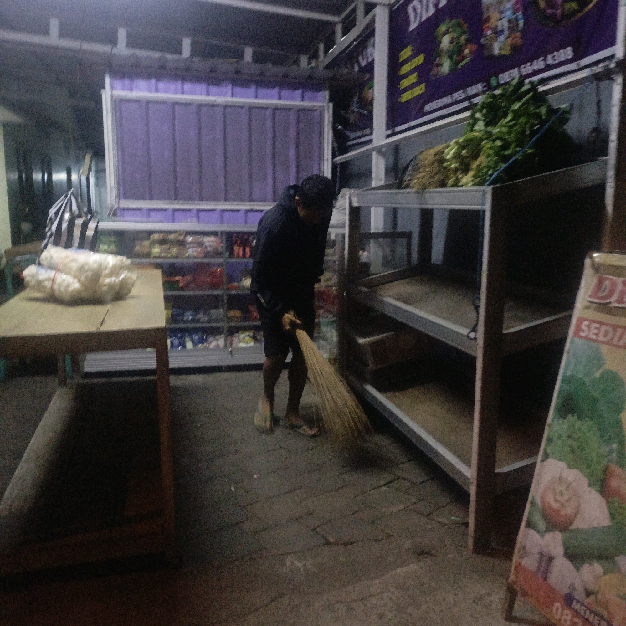 A person wearing a black hooded jacket and black shorts is sweeping a brick floor with a broom. The person is facing away from the camera. To the left of the person is a wooden table with packages of white food items wrapped in plastic and tied with red string. Behind the table and to the left are shelves filled with various packaged goods. To the right of the person are metal shelves, some of which are empty and others contain some items. A large sign with purple lettering and images of vegetables is visible in the background, to the upper right. A small portion of a sign with red and green colors and the text "SEDIA" is visible in the lower right corner. The scene appears to be an outdoor market or a small shop. The lighting suggests it might be evening or indoors. The location is in the city of Salatiga, Indonesia, likely at a market stall.