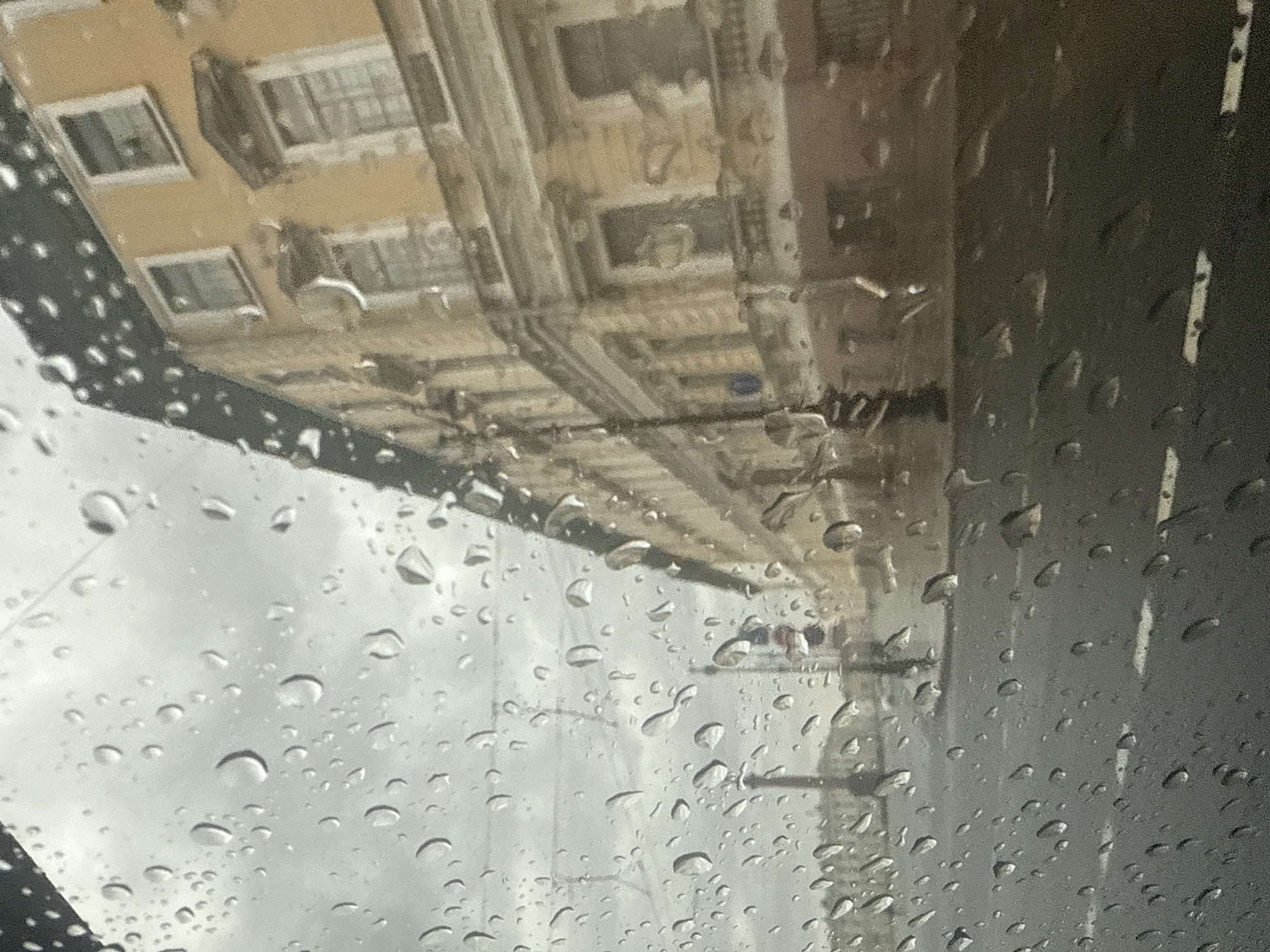 The image shows a street view in Saint Petersburg, Russia, observed through a window covered in raindrops. A yellow building with multiple windows and ornate architectural details lines the street. The sky is overcast and gray. A traffic light is visible in the distance, and the road surface appears wet. The overall impression is of a rainy day in an urban environment.