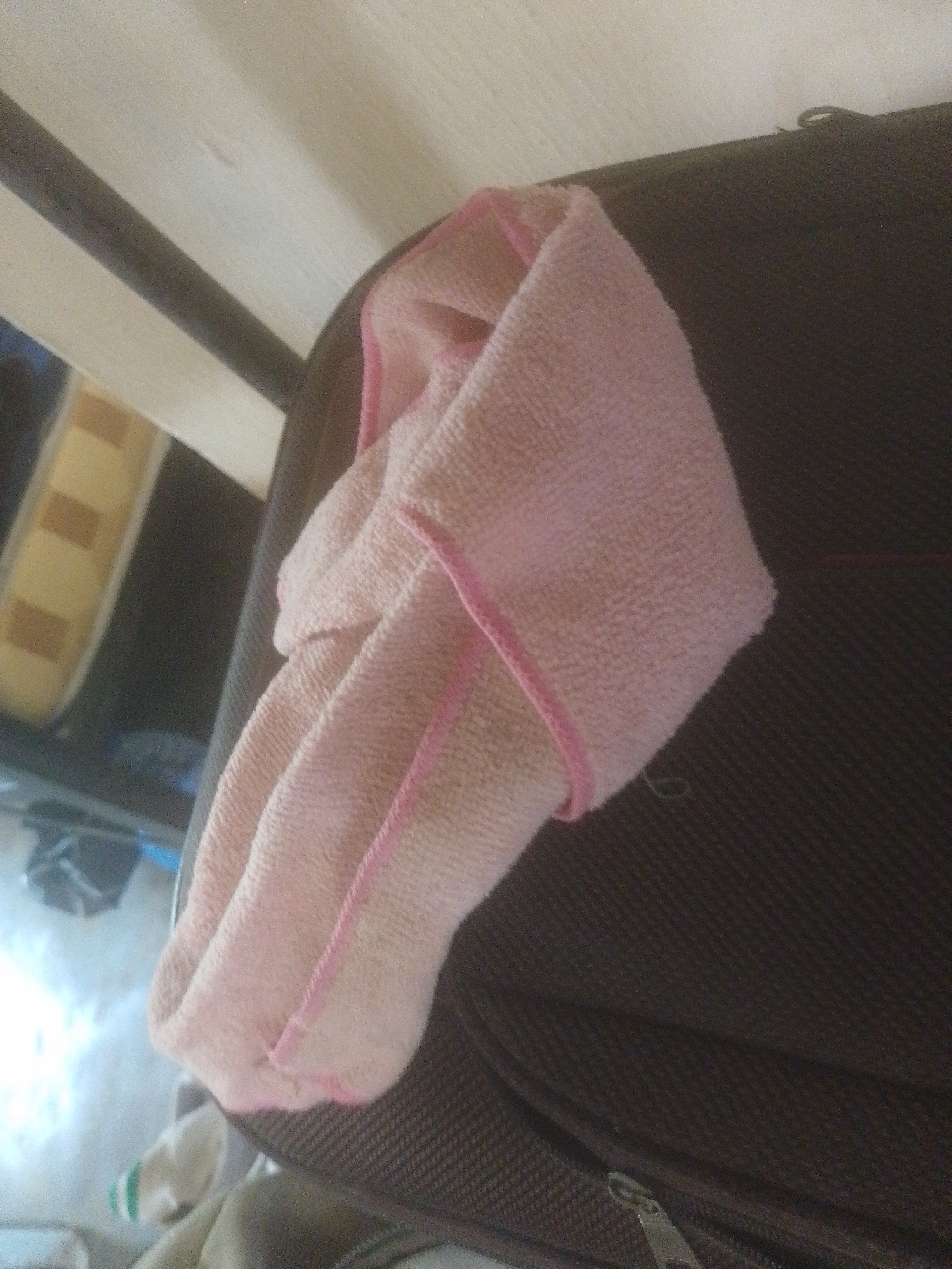 A pink microfiber cloth is draped over a dark brown, textured surface, possibly a suitcase. The cloth is folded and bunched up, with a pink edge binding visible. The background consists of a light-colored, slatted surface and a blurry area showing what appears to be a yellow and brown object with rectangular patterns. The scene is poorly lit and lacks clarity. No specific location in Danbazau, Nigeria, can be determined from the visual information provided.