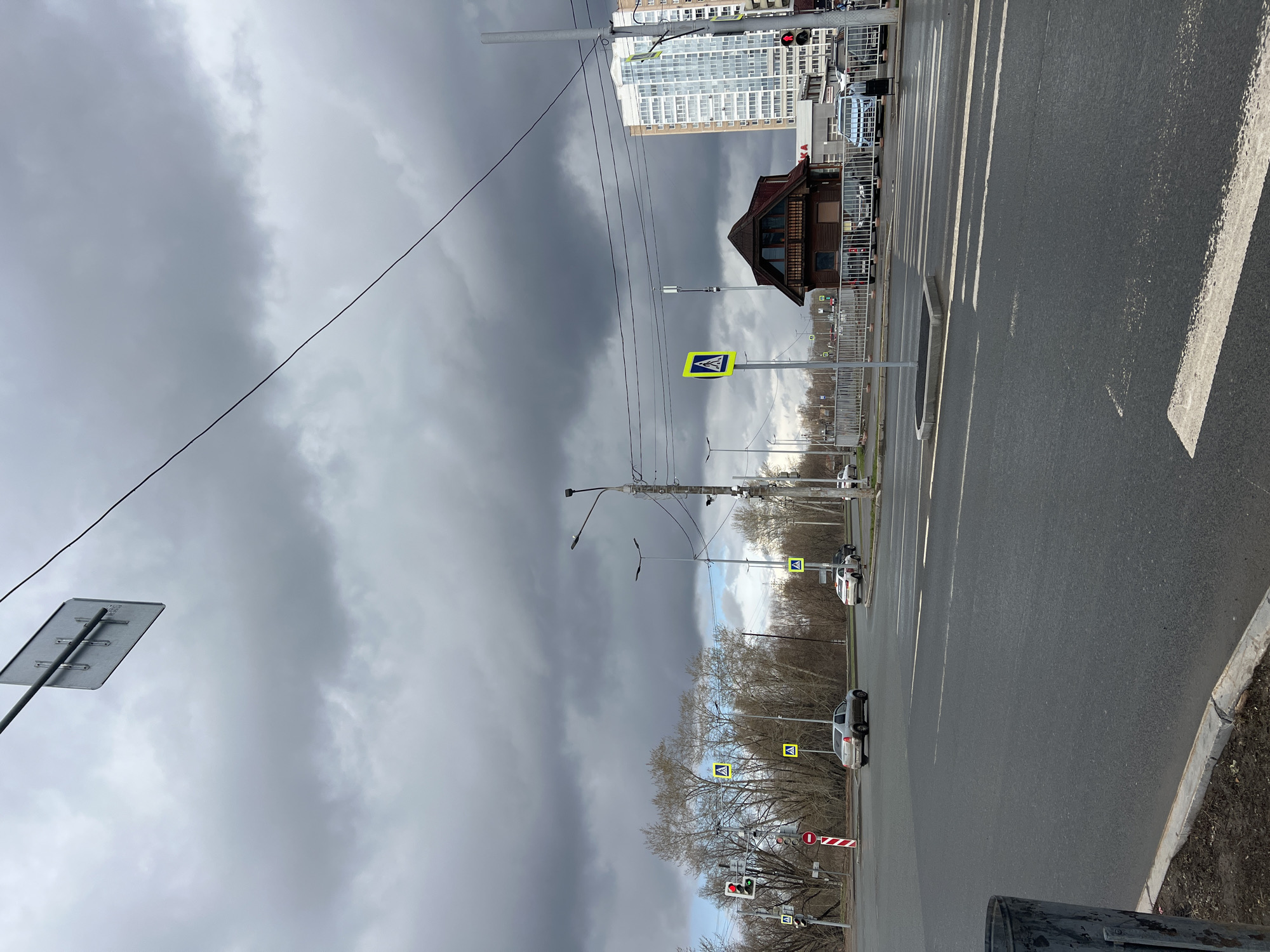 The image depicts a street scene in an urban or suburban environment, likely in Perm, Russia, under a heavily overcast sky. The photograph is taken from a slightly elevated perspective, looking down a multi-lane asphalt road.
The sky is dominated by dark, heavy grey clouds, suggesting an impending storm or a very dull day. Overhead, numerous utility lines crisscross the frame.
On the right side of the road, a wide pedestrian crosswalk (zebra crossing) is clearly marked with white stripes. Further down the road, additional crosswalks are indicated by yellow square signs with black pedestrian symbols. Several cars, including white and grey sedans, are visible on the road, some appearing to be stopped at a traffic light which displays a red signal on the right side of the frame. Another traffic light further down the street shows a green signal. A red circular sign with a white horizontal bar, typically indicating "No Entry" or "Stop," is also present.
Along the right side of the road, a metal fence separates the roadway from a sidewalk or undeveloped strip. Trees with bare branches line the street, suggesting either late winter or early spring. In the background, a modern, tall building with numerous windows stands prominently, while closer to the foreground, a distinctive two-story wooden building with a peaked roof and multiple windows adds an architectural contrast. On the side of the modern building, a faint sign, possibly reading "A-KA" or "AKB," is visible. Streetlights and various poles for traffic signals and utility lines are scattered throughout the scene. No people are visible.