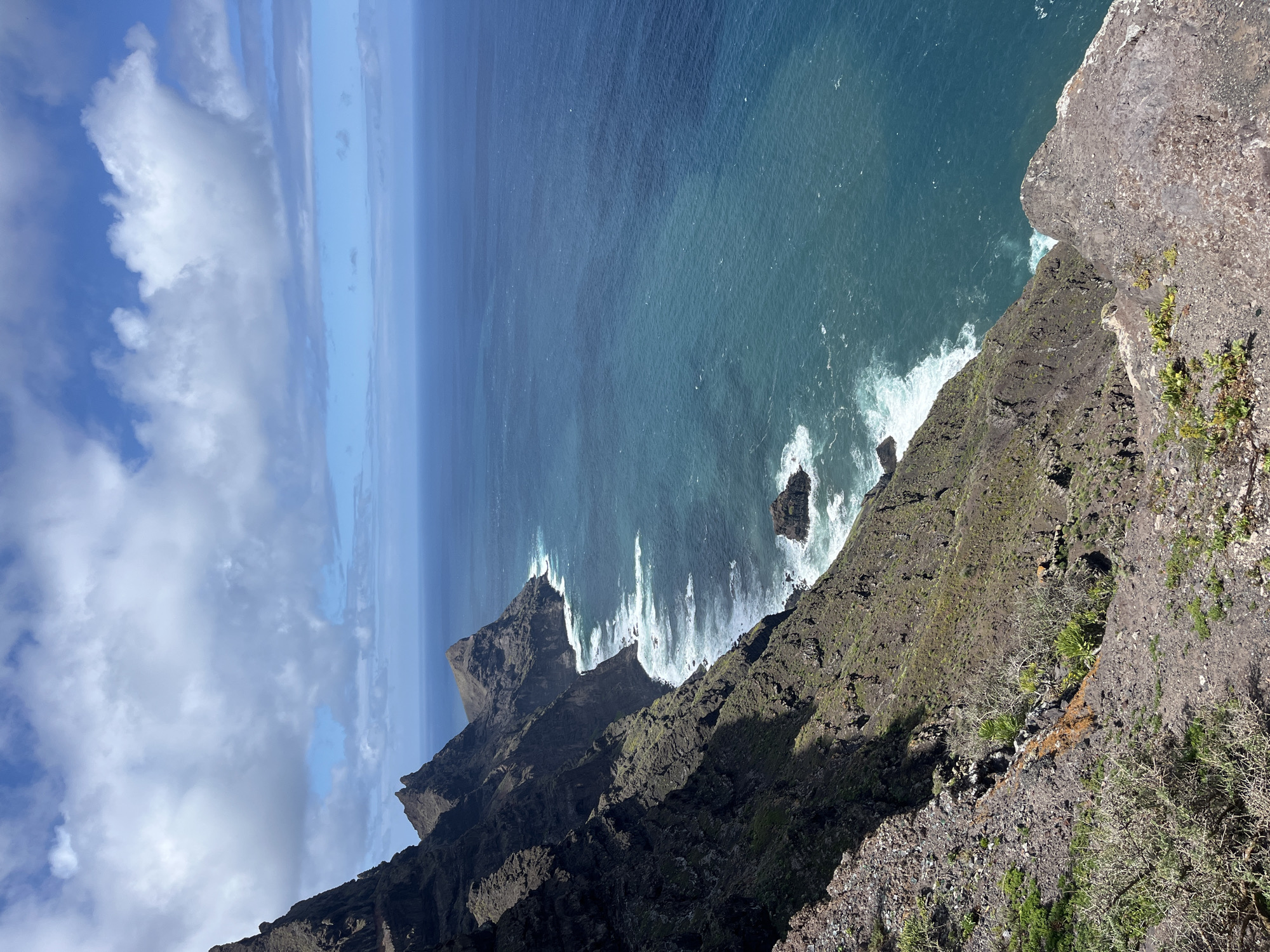 A high-angle shot depicts a rugged coastline in Agaete, Spain, with steep cliffs plunging into the Atlantic Ocean. The cliffs are primarily rocky and covered with sparse vegetation. Waves crash against the rocks below, creating white foam. In the distance, the ocean transitions from deep blue to a lighter shade near the horizon, which blends with the sky. The sky is partly cloudy with patches of blue. No people are visible in the image.