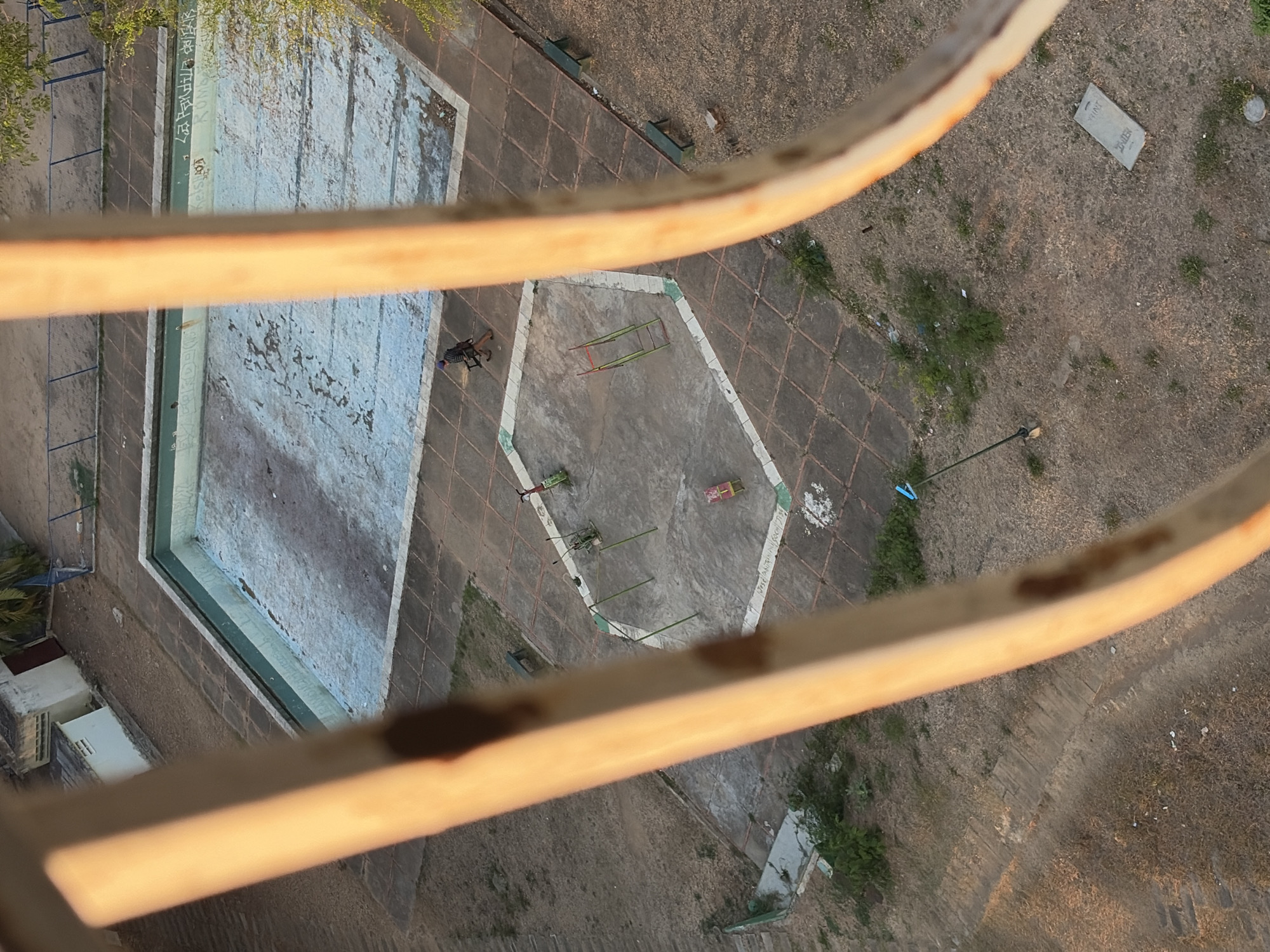 The image shows an aerial view of a derelict area in Ciudad Guayana, Venezuela. An empty swimming pool with faded paint is visible on the left. In the center, there is a hexagonal paved area containing abandoned playground equipment. A person is walking nearby. The surrounding ground is overgrown with sparse vegetation. The image is taken through an orange railing.