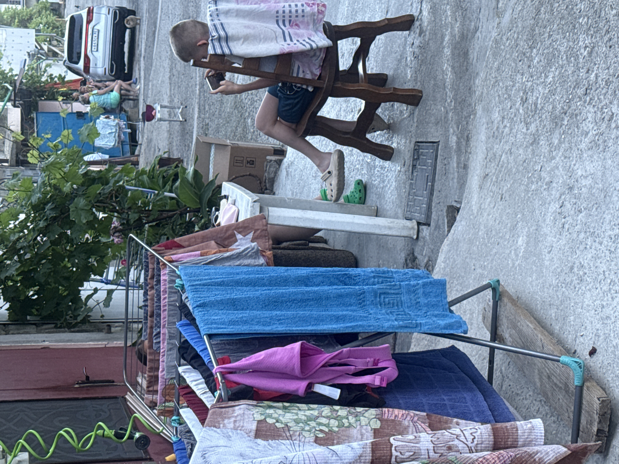 A young boy, seen from the back, sits on a wooden chair with a blanket draped over its back, engrossed in a smartphone. He is wearing dark blue shorts and a light-colored t-shirt. His legs are crossed, and his feet are bare. Beside him, a young child in bright green Crocs is also seated, though their face is not visible. In the foreground, a clothes drying rack is filled with various garments, including a bright blue towel, a pink garment, and patterned fabrics. To the left, a red door and a green coiled hose are visible. In the background, a white SUV is parked, and several people are seated on a bench, conversing. A small dog stands near the car. The scene appears to be an outdoor residential area, possibly a courtyard or street, with a mix of urban elements and vegetation.