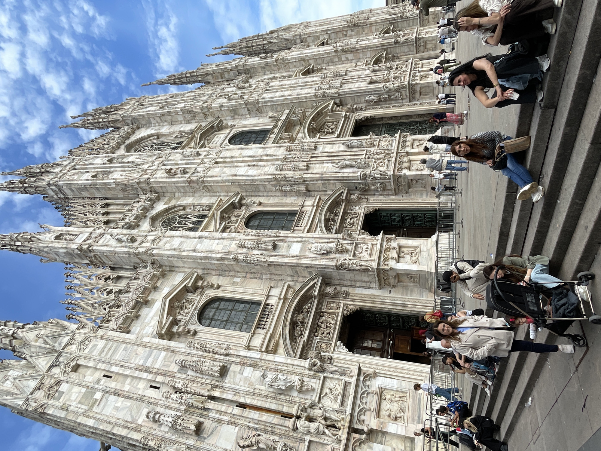 The image captures the façade of the Milan Cathedral (Duomo di Milano) in Milan, Italy, from a slightly angled perspective. The building's elaborate Gothic architecture, marked by its white marble exterior, pointed arches, numerous statues, and spires, extends upwards towards a blue sky with scattered clouds. In the foreground, a set of stone steps lead up to the cathedral's entrance. Several individuals are present on the steps. Some are seated, and others are standing or walking. They include adults. Interactions appear casual, with people potentially taking photos or simply resting. The environment suggests a busy tourist location.