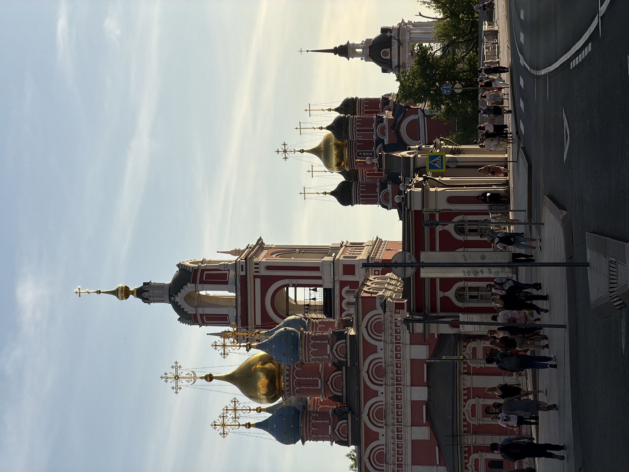 The image depicts a vibrant urban scene in Moscow, Russia, featuring a section of a street adjacent to a Russian Orthodox church.
**Buildings:** The architectural style of the church is distinct, with red and white facades, and multiple onion-shaped domes with golden or blue finishes and prominent crosses on top. It is adorned with numerous windows and architectural details. Another structure, also likely part of the church complex, stands further back, with a different architectural style and a dark roof.
**People:** A group of people is walking along the sidewalk next to the church. They appear to be diverse in clothing and age, potentially tourists or locals going about their day.
**Street:** The street is partially visible on the right side of the image, with painted white lines. A pedestrian crosswalk sign is seen near the curb.
**Sky:** The sky is light blue with some clouds, suggesting it could be either early morning or late afternoon.
**Overall Impression:** The scene conveys a sense of bustling urban life juxtaposed with the historical and religious significance of the church. The image appears to be taken during daylight hours. The specific location in Moscow is difficult to determine without more information, but the architectural style is typical of Russian Orthodox churches found throughout the city.
