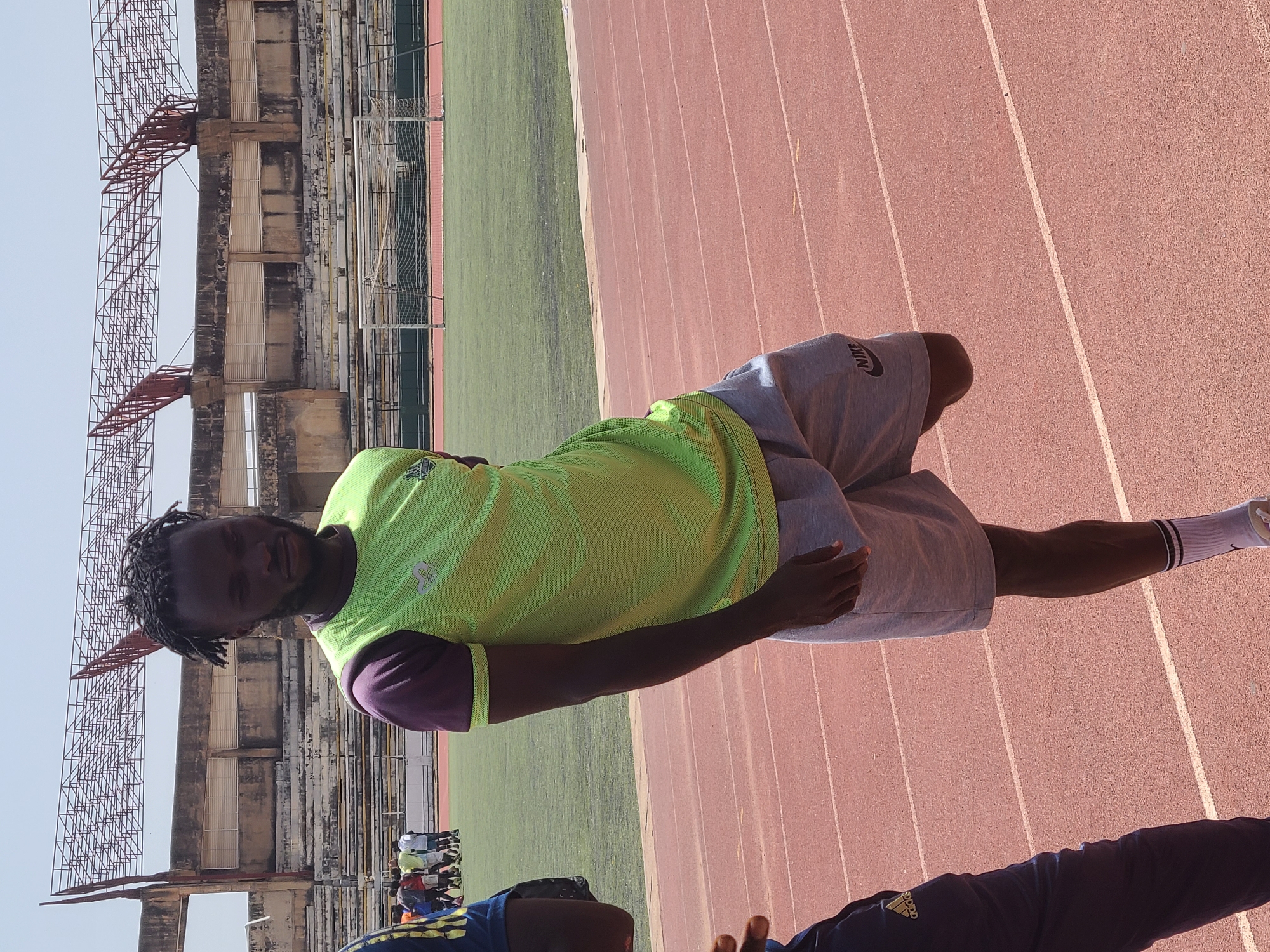 A man stands on a red running track next to a green football field at the New Stadium in Nigeria. The stadium structure is visible in the background. The man is wearing a neon green shirt with purple sleeves and grey shorts. He has dreadlocks. Another person wearing a dark blue tracksuit is partially visible in the foreground. A group of people are gathered in the background. The sky is light blue.