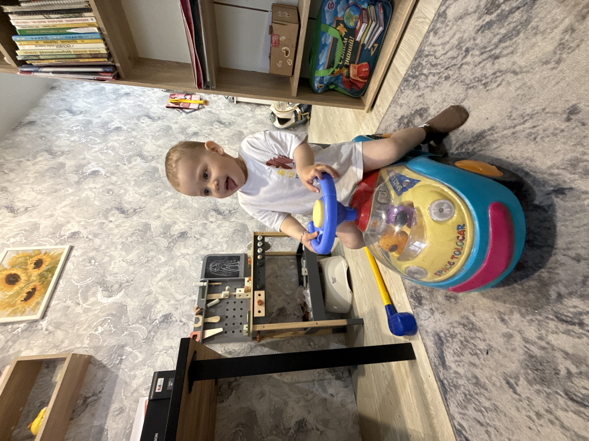 The image captures a child playing with a ride-on toy. The child, dressed in a white t-shirt, sits on a multi-colored toy car with a blue steering wheel. The toy car has "SPACE TOLOCAR" printed on its side and includes a toy golf club leaning against it.
The setting appears to be a room with wallpaper featuring a gray and white marbled pattern. The room includes shelves filled with books and objects, and a painting of sunflowers hangs on the wall. A small workbench with tools is also visible.
No specific location is identifiable within the city of Rusanovo, Russia, based on this image.