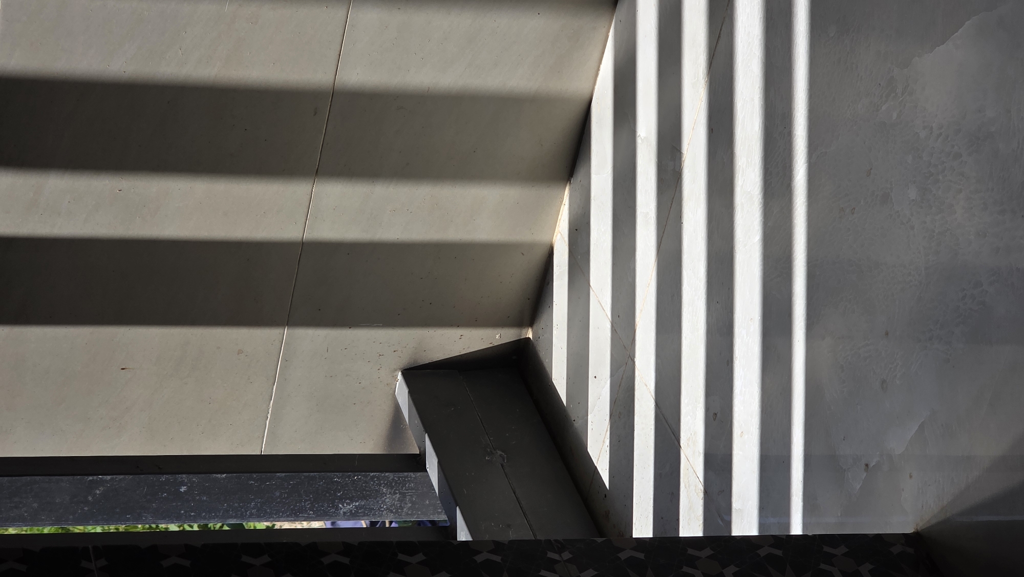 The image shows a corner of a tiled floor and wall in Bac Natural Heritage Area Trang, Vietnam. Sunlight creates stark shadows of parallel lines. A dark gray metal frame is visible at the bottom of the frame with hints of vegetation. The tiles appear to be of two different colors/textures.