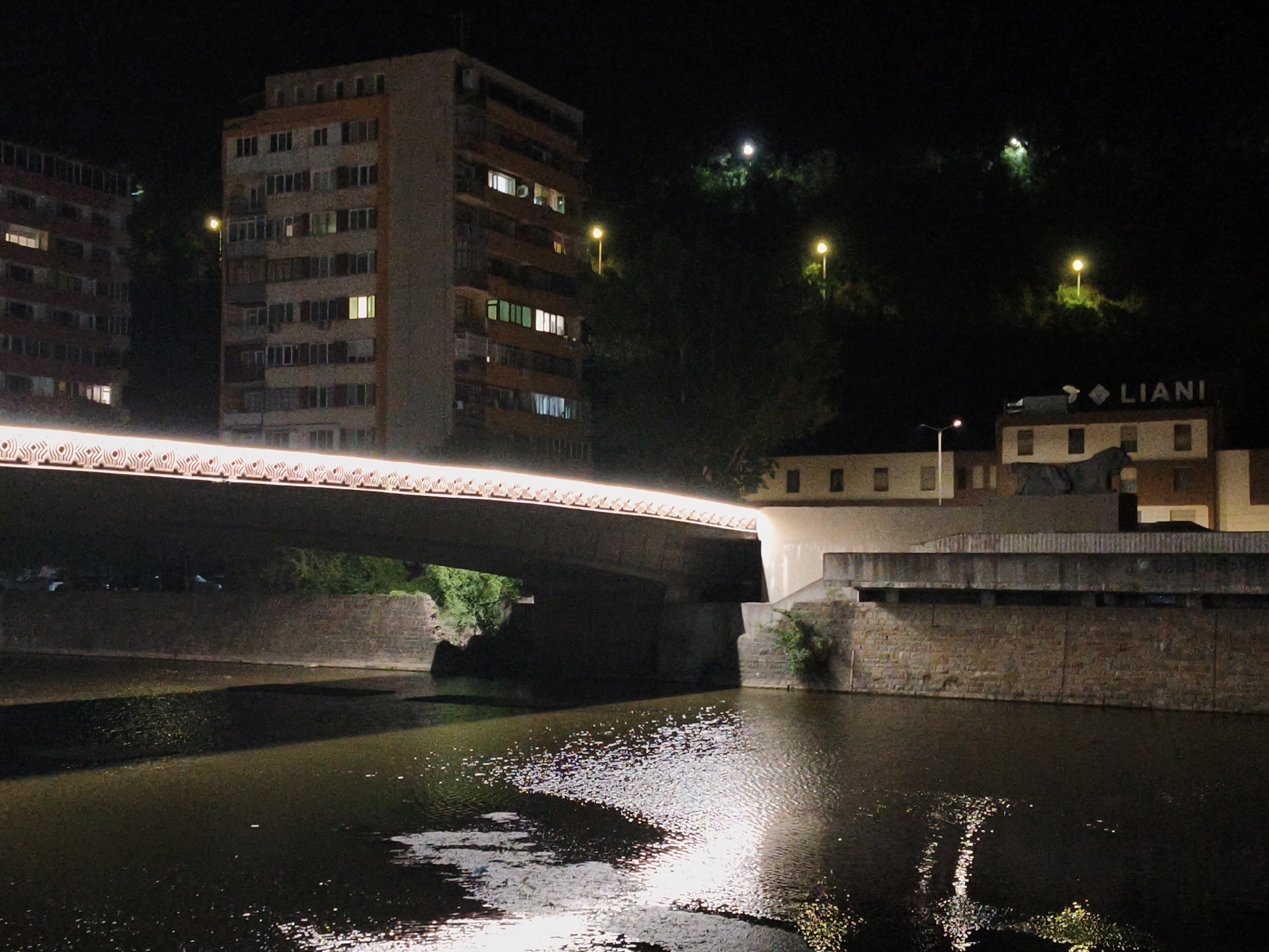 The image depicts a nighttime scene in the city of Lovech, Bulgaria, focusing on the covered pedestrian bridge over the Osam River. The bridge is illuminated from above by a continuous strip of warm-toned lights. The river below reflects the light from the bridge and the surrounding urban environment, creating shimmering patterns on its surface. On the left bank of the river, a multi-story residential building with many illuminated windows is visible. On the right bank, another building with a sign reading "LIANI" is present, along with a stone embankment and vegetation. The background shows a dark, forested hillside with several streetlights illuminating parts of the terrain. The overall atmosphere is nocturnal and serene, highlighting the urban infrastructure and natural elements of Lovech.
