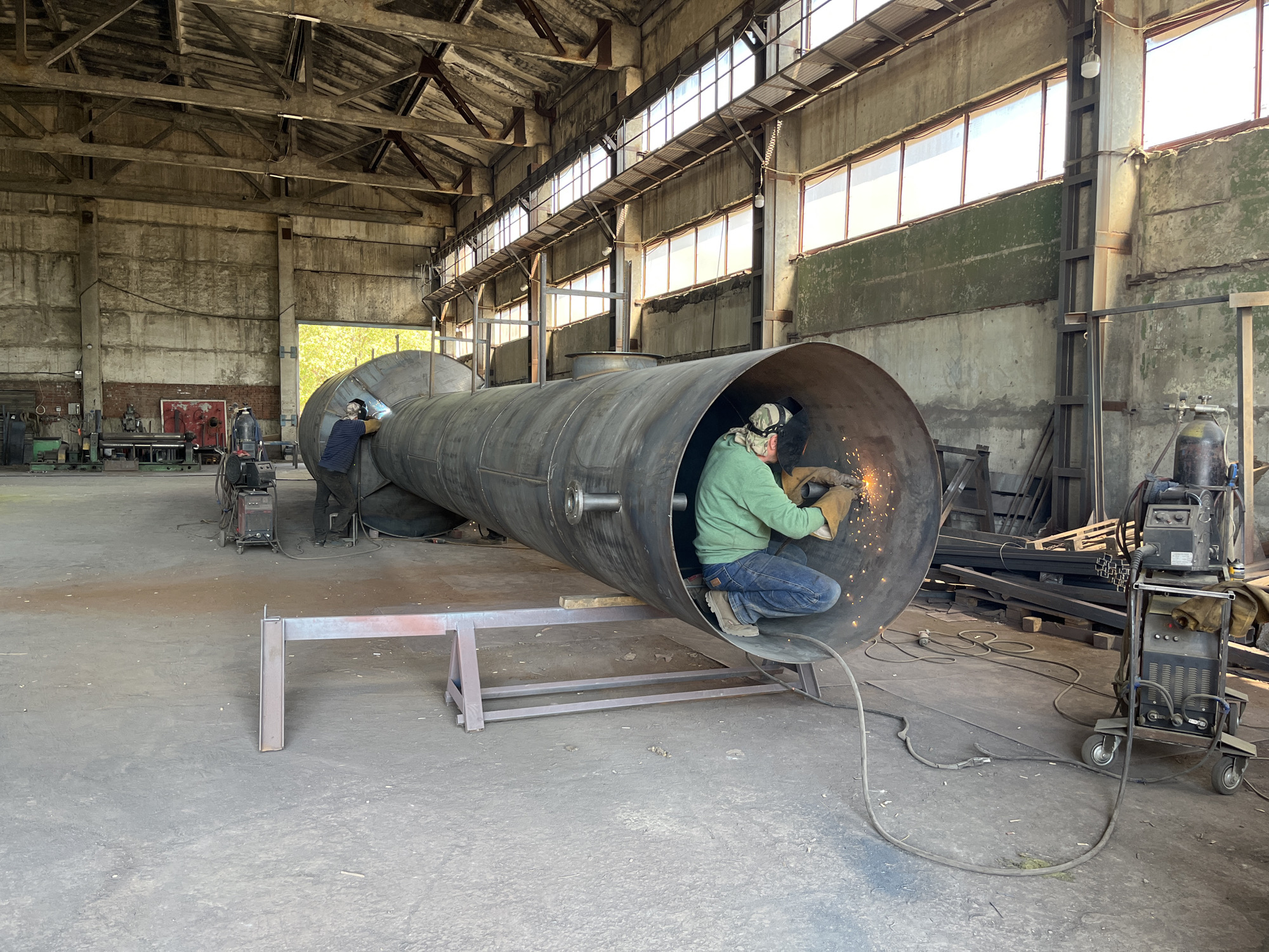 This image depicts an indoor industrial workshop setting, likely a fabrication or repair facility, located in Taganrog City District, Russia. The environment is characterized by a large, open-plan space with a high, exposed-beam ceiling and concrete walls. Natural light streams in from numerous windows along the upper section of the building.
The primary focus is a large, cylindrical metal tank lying horizontally on the floor, supported by metal stands. Two individuals are actively engaged in welding work on this tank. One welder, wearing a green hooded sweatshirt, jeans, and protective gear including a welding mask and gloves, is positioned inside the opening of the tank, sparks flying as they work. Another welder, also in protective attire, is seen near the left end of the tank.
Various industrial equipment is visible in the background, including welding machines on wheeled carts, metal stock, and other machinery, suggesting ongoing construction or maintenance activities. The floor is concrete, showing signs of industrial use with dust and some scattered debris. The overall scene conveys a working environment where heavy fabrication is taking place.