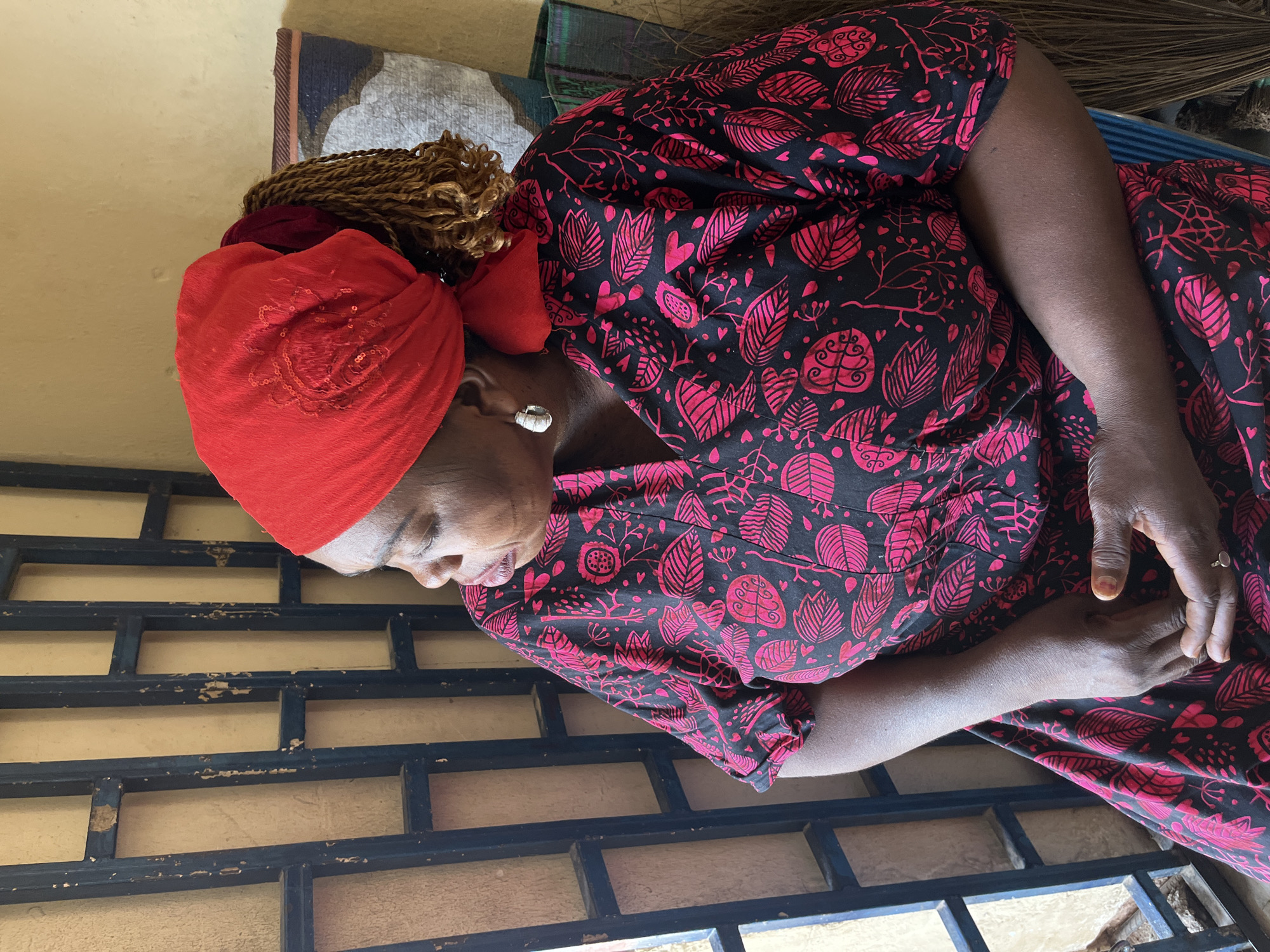 A close-up, mid-shot image captures a woman from the chest up, positioned slightly to the right, with her head turned downwards and to the left. She has dark skin and wears a bright red head wrap, under which light brown braided hair is visible near her left ear. A single silver stud earring adorns her left ear. She is dressed in a black garment, likely a dress or top, intricately patterned with vibrant pink leaves, hearts, and abstract organic shapes. A silver ring is visible on the ring finger of her right hand, which rests gently, partially obscured by the fabric.
The woman appears to be in a state of quiet contemplation or rest, her eyes partially closed or cast downwards. The lighting suggests it is daytime, with diffused natural light illuminating the scene.
The background elements include a light yellow, textured wall to her left. Below her, occupying the lower third of the frame, is a dark, grid-like structure, possibly a window or security grill, showing signs of wear with peeling paint. Behind her, to her right, various textured fabrics or mats in blues, greys, and browns are visible, alongside a bundle of thin wooden sticks, resembling a broom. These details suggest an indoor or semi-outdoor domestic setting in a lived-in environment. There is no visible text in the image.