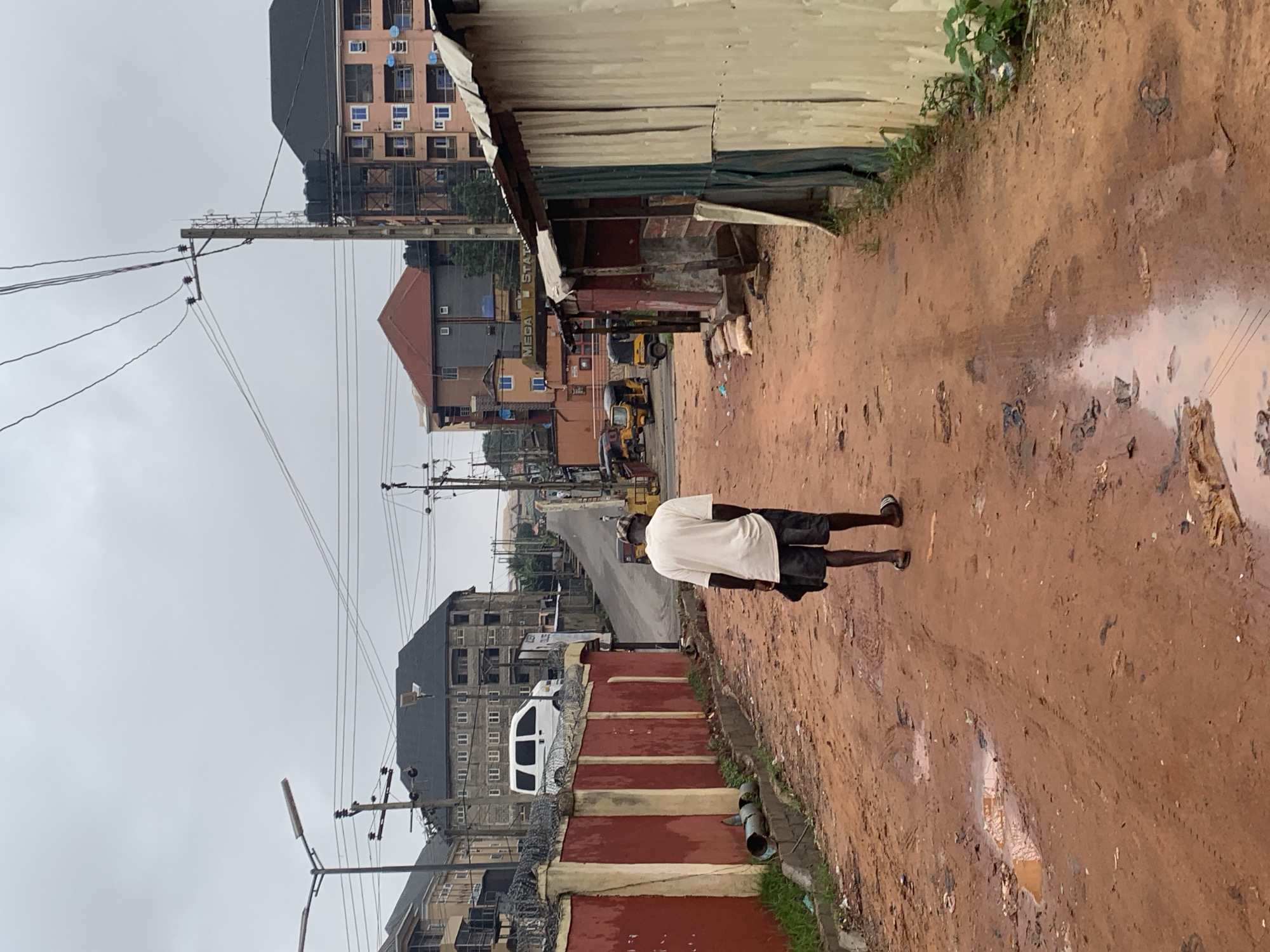 A lone man wearing a white t-shirt and dark shorts walks away from the camera down a dirt road in Nkpor, Nigeria. He carries a dark bag over his right shoulder. The road is muddy with several puddles and tire tracks. On the left side of the road is a red concrete wall topped with barbed wire, with a white van parked behind it. Beyond the wall are multi-story buildings. On the right side of the road are several corrugated metal structures. In the background, the road slopes upwards towards more buildings and power lines crisscross the overcast sky. Three yellow and black auto-rickshaws are visible further up the road.