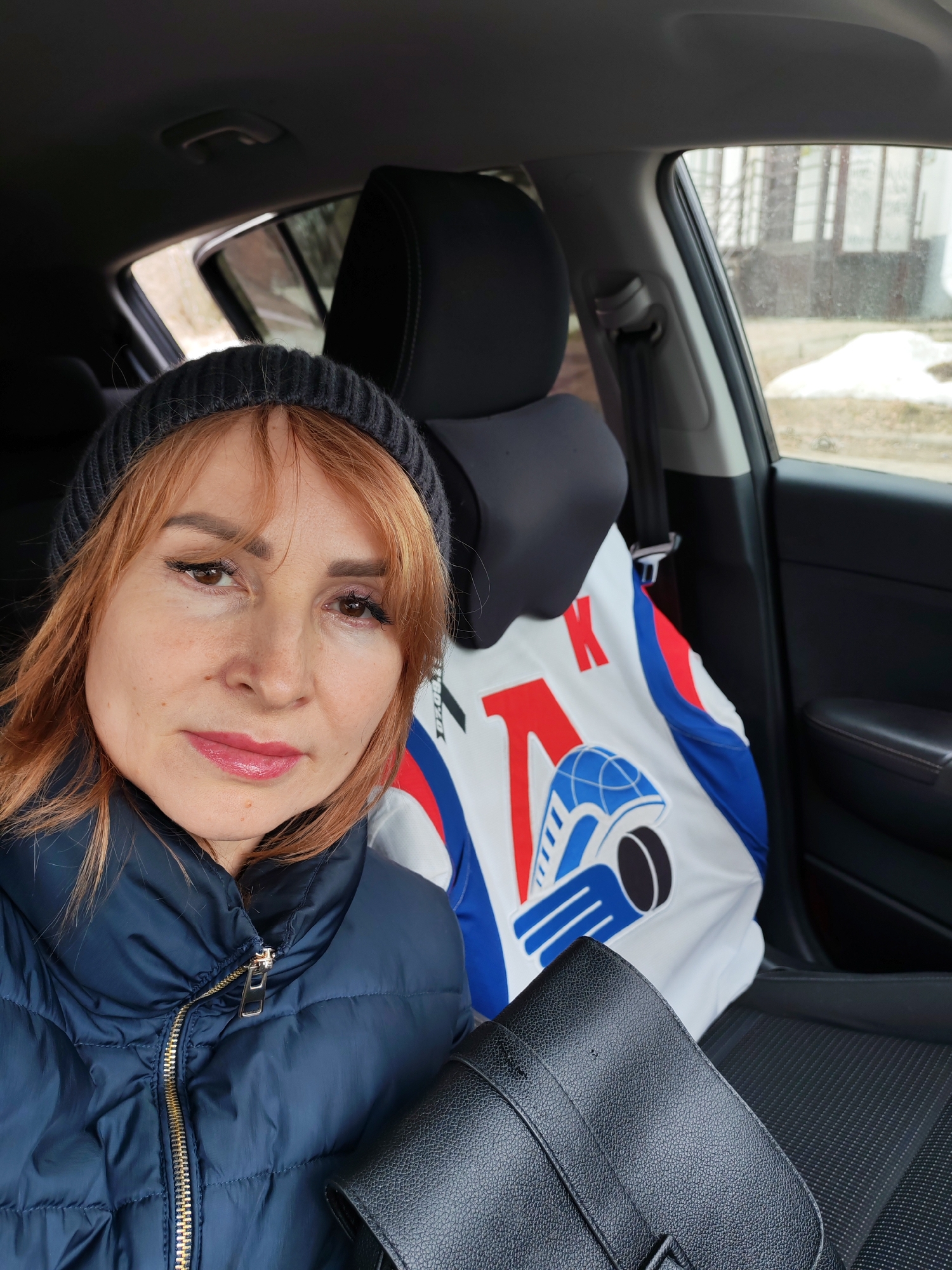 A woman with reddish-orange hair and a dark gray knitted hat is seated in the driver's seat of a car. She is wearing a navy blue puffer jacket. A black leather purse is visible in her lap. Behind her, hanging on the back of the passenger seat, is a white jersey with red and blue stripes. The jersey has a logo on it featuring a globe, a hockey puck, and a stylized image of hands. Through the car window, a blurred view of a building and patches of snow on the ground are visible. The location is identified as Chaikovsky, Russia.