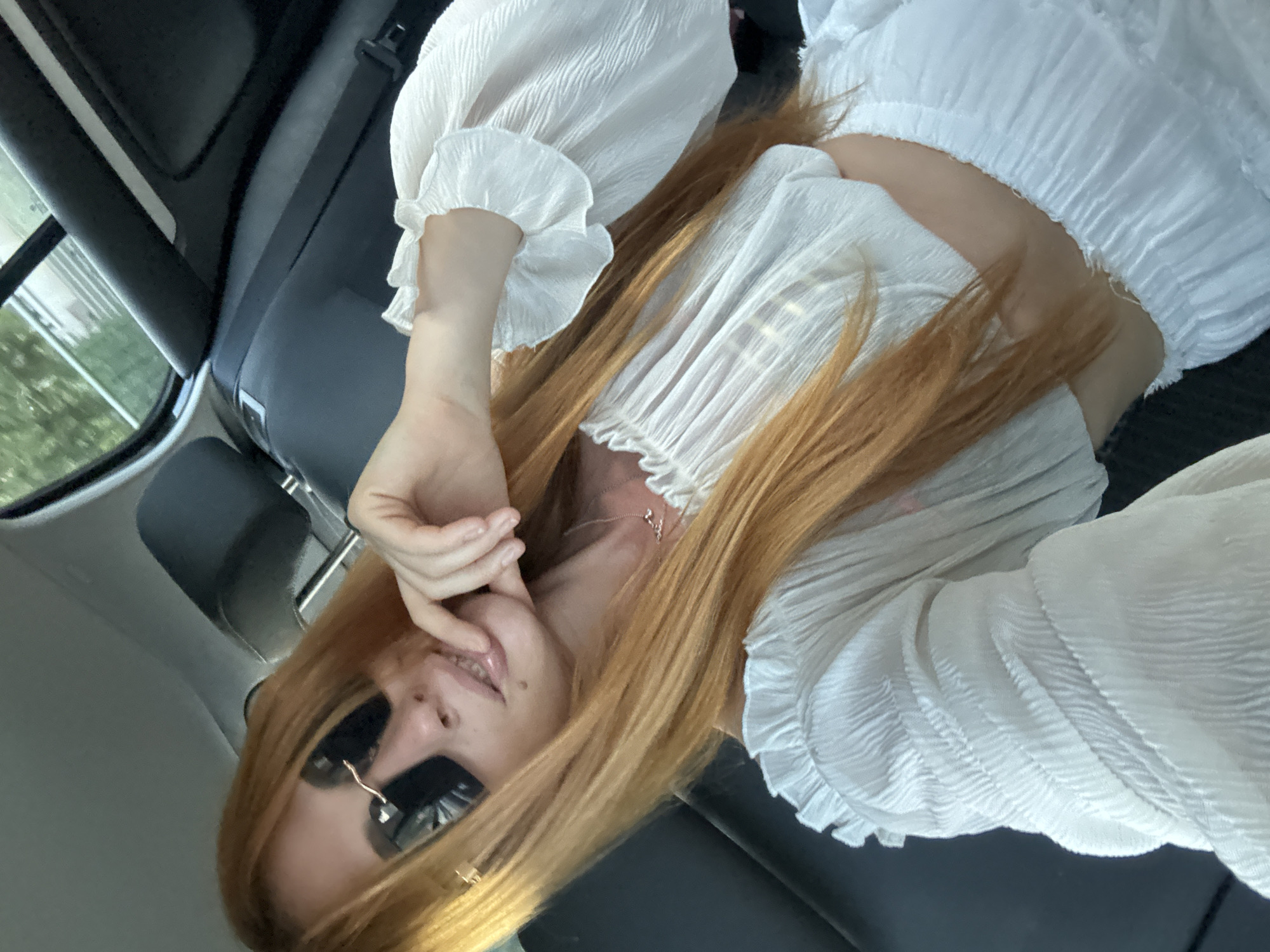 A female individual is depicted in the interior of a vehicle, positioned to capture a selfie. She has long, straight, reddish-blonde hair and wears dark, rectangular sunglasses with visible metal accents. Her lips are slightly parted, revealing teeth, and a small dark mark is present on the lower right side of her chin. A delicate silver-colored necklace adorns her neck. She is dressed in a white, textured, off-the-shoulder crop top featuring ruffles at the neckline and cuffs, paired with a matching textured skirt, parts of which are visible. Her right hand is raised, with fingers lightly touching her chin. The car interior features dark grey/black upholstery for the headrest and door panels, a light grey headliner, and a visible seatbelt buckle on the right. Through the car window, blurred green foliage and indistinct light-colored structures are visible, indicating an outdoor setting under strong natural light. The scene is located in Petrovac na Moru, Montenegro.