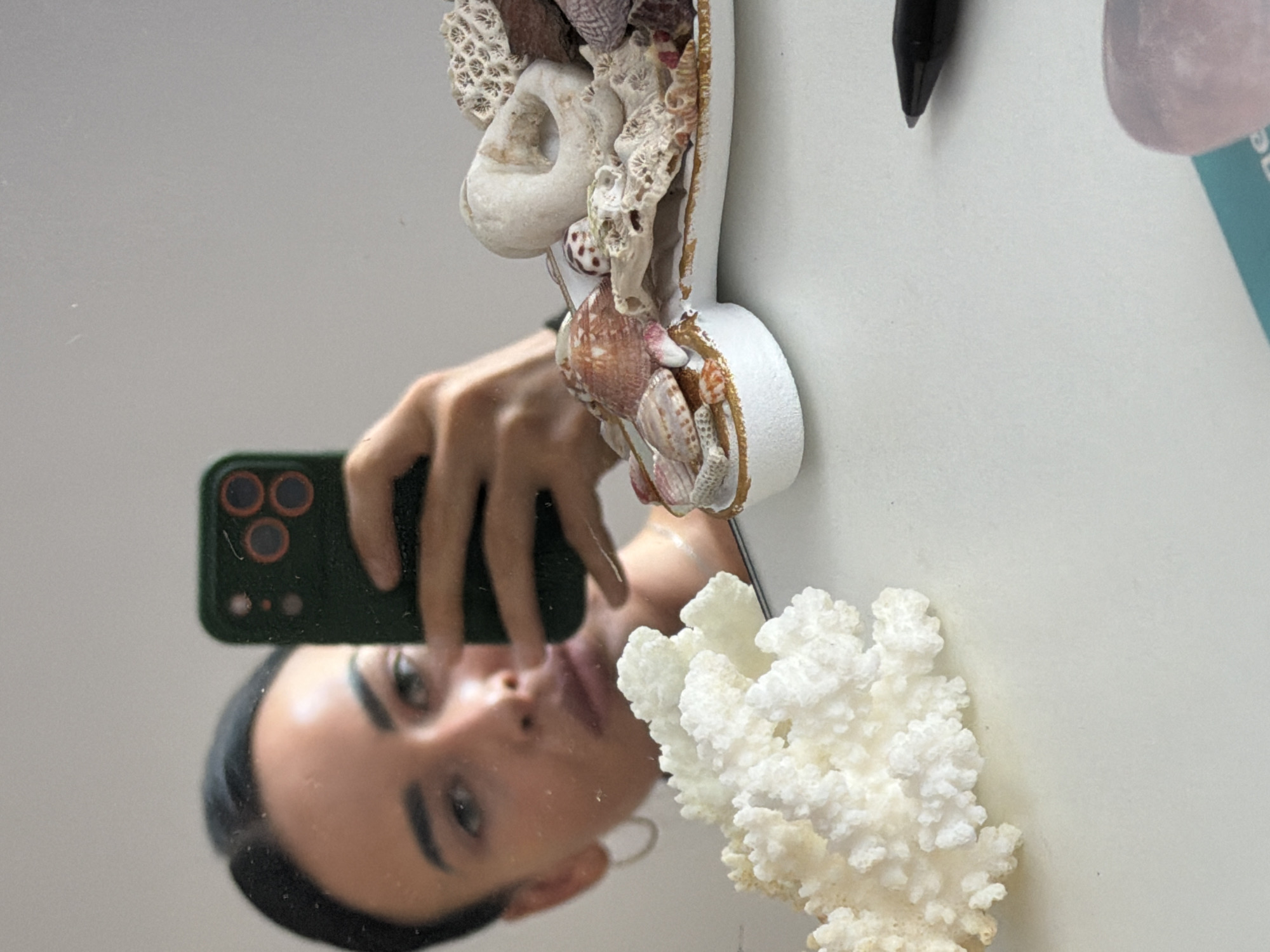 The image displays a mirror selfie, captured with the camera rotated 90 degrees clockwise from standard vertical orientation. In the reflection, a person's face is visible in the lower-left, looking directly into the camera. They are holding a dark green smartphone with a prominent triple-lens camera system and a smaller sensor/flash array on its back.
The rest of the frame is filled with an intricate display of marine-themed decorations. A large, textured piece of white coral occupies the bottom-right corner. Above this, and extending towards the center, is a dense arrangement of various seashells and coral fragments in shades of white, beige, brown, and pink, mounted on a white base. These include several types of spiral and bivalve shells, along with branching and porous coral pieces. Towards the upper right, a black pen or stylus points downwards, and a light pink, translucent object (possibly a crystal or polished stone) is partially visible, resting next to a small, turquoise-colored card with illegible, blurred text. The background behind these objects is a plain, light-colored surface, suggesting an indoor setting like a shelf or desk. The person in the reflection has a neutral expression.