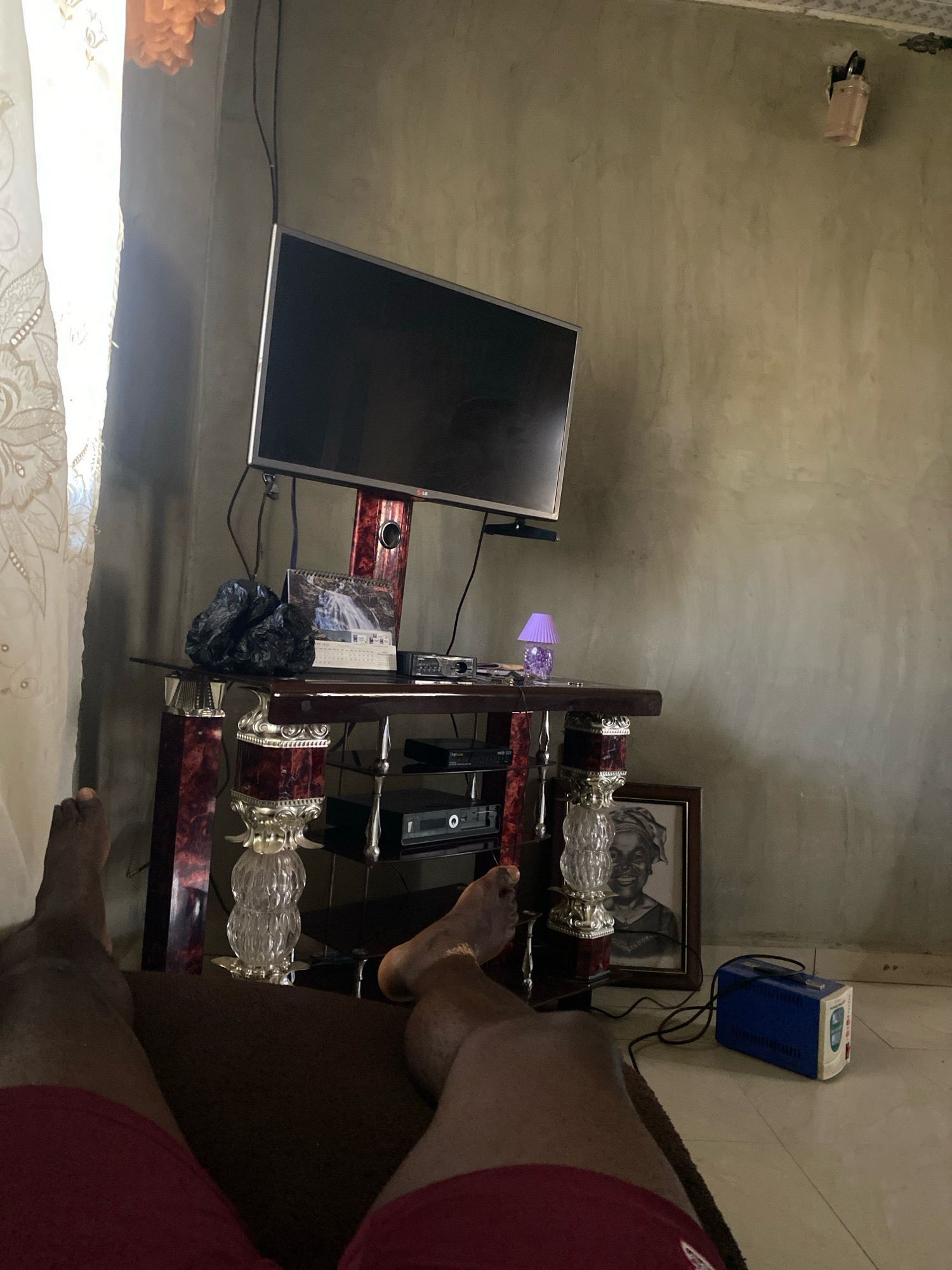 In an indoor setting, likely a living room in Jos, Nigeria, a person's legs are visible in the foreground, resting on a dark brown sofa. The person is wearing red shorts. The room features a television set mounted on a dark wood and glass entertainment unit. On the unit, there are various items including a calendar depicting a waterfall, a black object wrapped in plastic, a small purple lamp, and other electronic devices. To the right of the entertainment unit, a framed black and white portrait of an elderly woman is leaning against the wall. On the tiled floor, a blue and white voltage stabilizer is connected to a black wire. The walls are a muted, textured gray. A white curtain with an intricate pattern is visible on the left side of the frame, with a glimpse of an orange decorative item hanging above it.