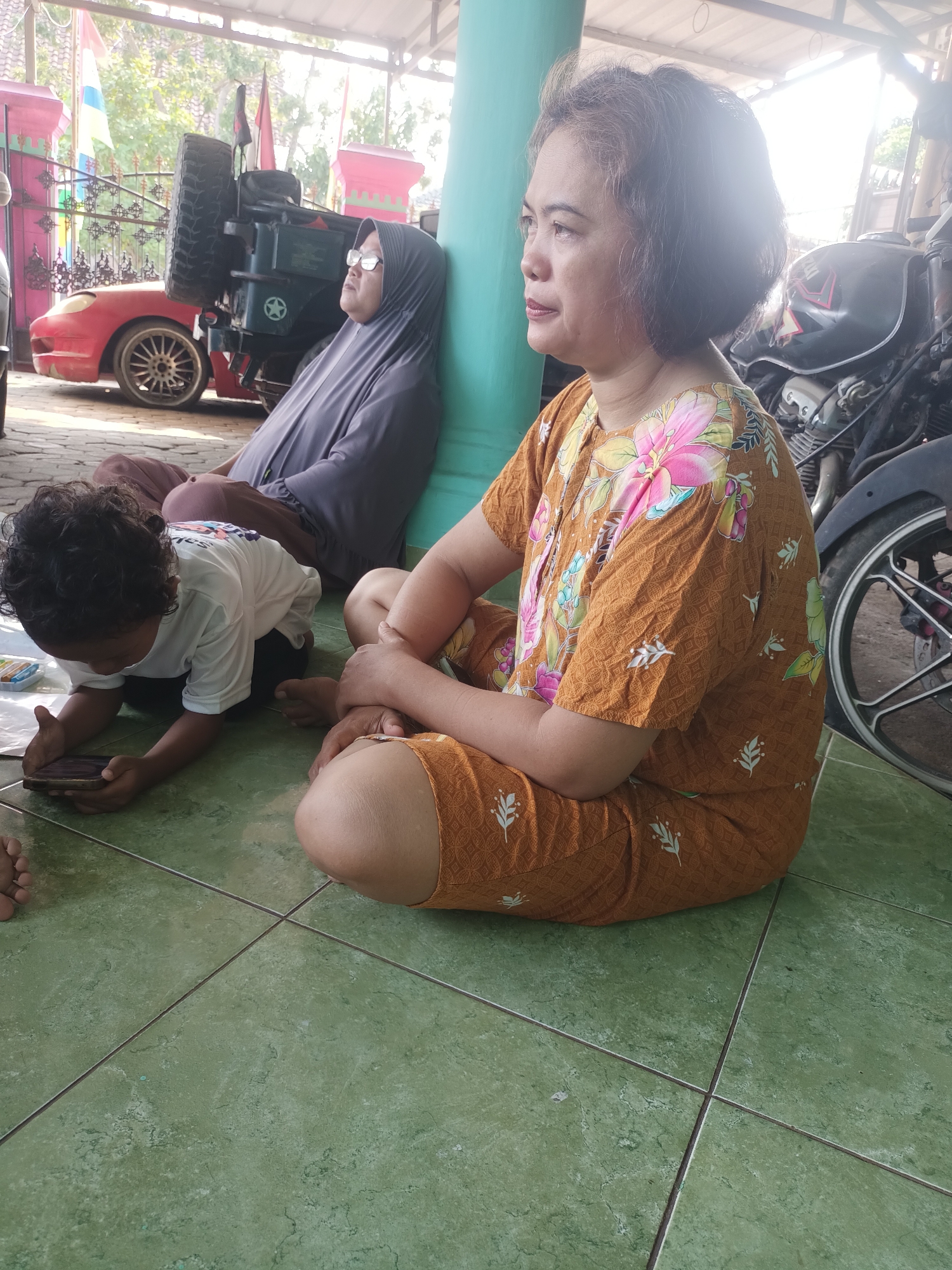 The image depicts three individuals seated on a green tiled floor under a covered structure, possibly a patio or carport, in Metro, Indonesia. The individual in the foreground, a woman with short, dark hair, wears a brown garment with a floral pattern and is seated with her legs crossed. To her left, a child with curly dark hair, dressed in a white t-shirt, is engrossed in a mobile phone. Behind the child, a woman wearing a grey hijab and glasses leans against a teal-colored pillar, facing away from the camera. In the background, a red car and a dark-colored vehicle, possibly a motorcycle, are visible, along with some flags and structural elements of a building. The lighting suggests it is daytime, with sunlight filtering through the cover.