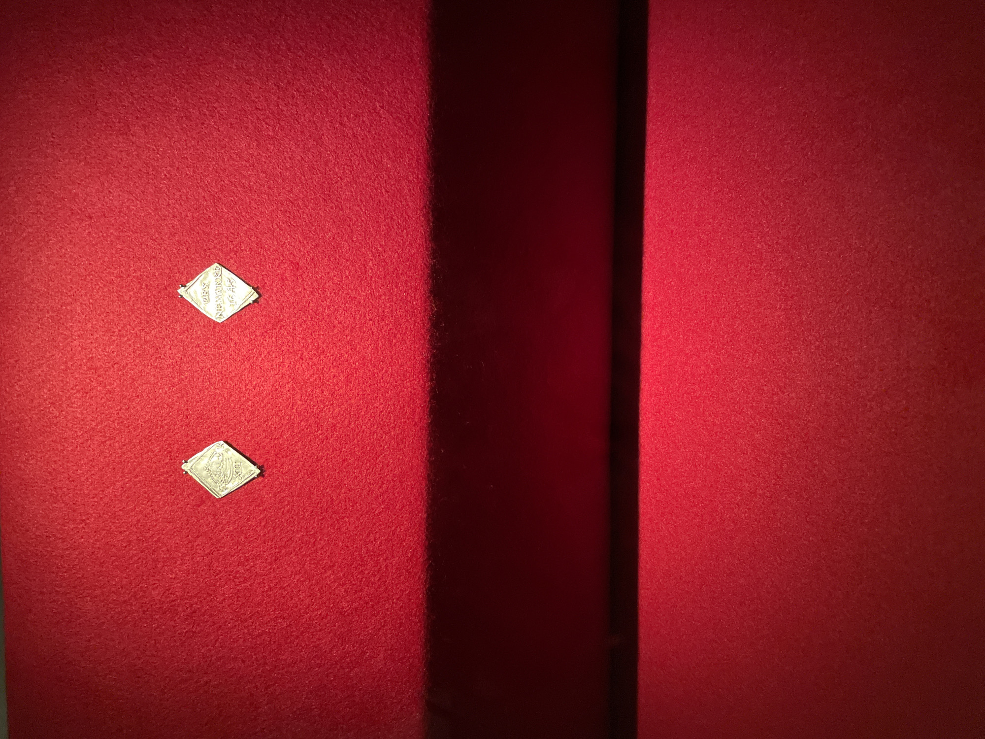The image features a close-up of what appears to be a red, possibly velvet or felt-covered surface with two small, diamond-shaped objects attached to it. The objects are silver or light-colored and seem to have inscriptions or designs on them. The background is a darker shade of red, possibly due to shadow or a change in the surface material. There are no discernible people, actions, or interactions in the image. Given the presence of “Barcelona” on the artifacts, the scene would likely be in the city of Barcelona, Spain.