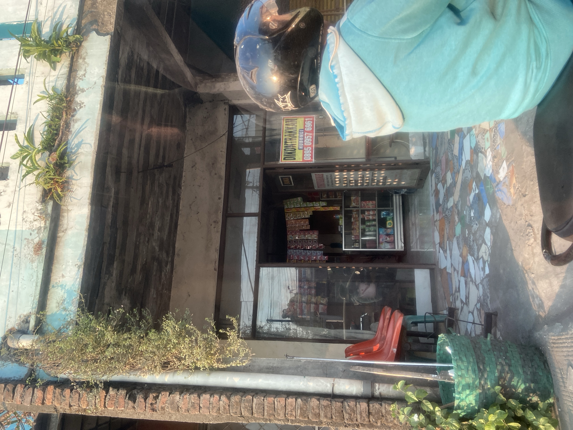 The image captures a street-level view of a shop front in Jekulo, Indonesia. Sunlight casts strong shadows across the scene. In the foreground, a person on a motorbike wears a black helmet and a turquoise hooded jacket. The shop itself has a glass entrance and displays various goods on shelves. A sign on the shop reads "DIKONTRAKKAN" with a phone number. Red plastic chairs are visible outside the shop entrance, and a large green woven basket sits to the left. The building's facade features peeling paint and vegetation growing from upper ledges. The ground in front of the shop is tiled with a mosaic pattern.