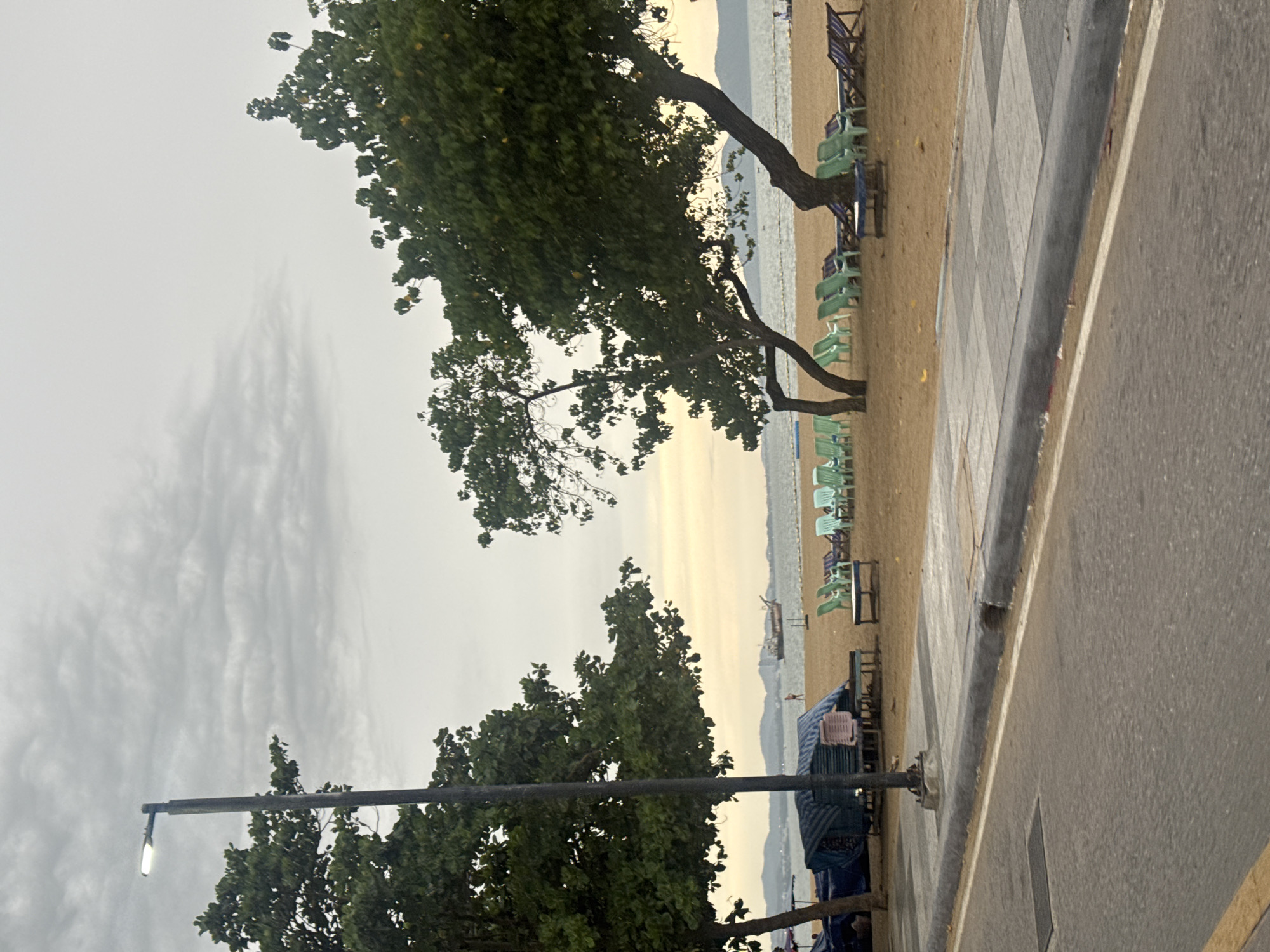The image depicts a beachside scene in Pattaya City, Thailand, under an overcast sky, suggesting it's either early morning or late afternoon. The foreground is dominated by a street with a paved sidewalk featuring a diamond pattern. Adjacent to the sidewalk is a sandy beach lined with rows of empty green and blue beach chairs, some neatly stacked. A large tree with dark bark and lush green foliage extends into the frame from the left, its branches reaching towards the sky. In the background, the calm sea stretches out, with a few boats visible on the horizon and hills forming a distant silhouette. There's a tall lamppost with a lit bulb on the left side of the image, partially obscured by trees. A temporary structure covered with blue and striped tarps, possibly a vendor's stall, is visible further down the beach, with a pink basket on top. The overall atmosphere is tranquil, with no people actively engaged in activities, hinting at a quiet moment before or after a busy period. The cloudy sky and muted colors contribute to a serene, somewhat subdued mood.